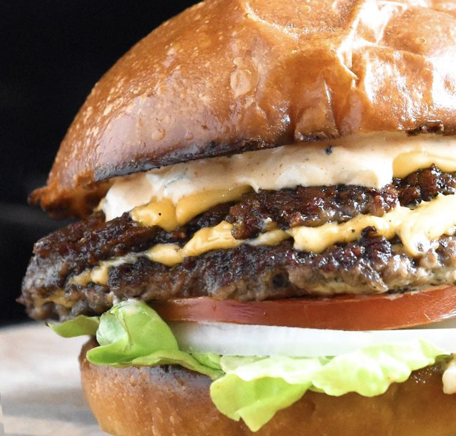 A close-up of a cheeseburger with a brioche bun, two grilled beef patties, melted cheese, sauce, lettuce, tomato, and onion.