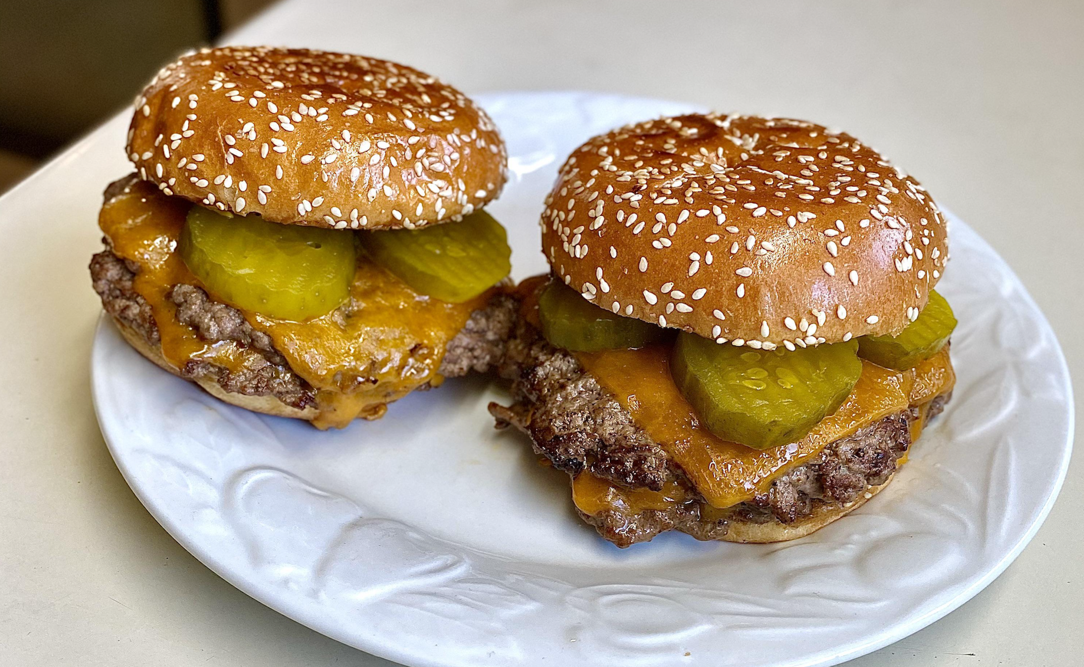 Two cheeseburgers with sesame seed buns, melted cheddar cheese, beef patties, and pickle slices on a white plate.