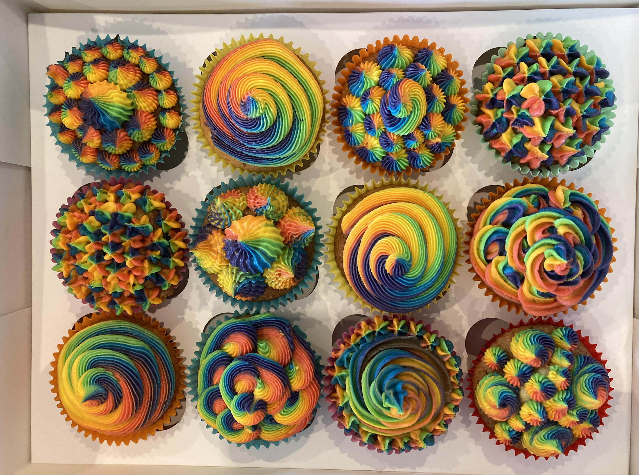 Twelve cupcakes with vibrant, rainbow-colored frosting arranged in a box, each decorated with a unique swirling or star-tipped pattern.