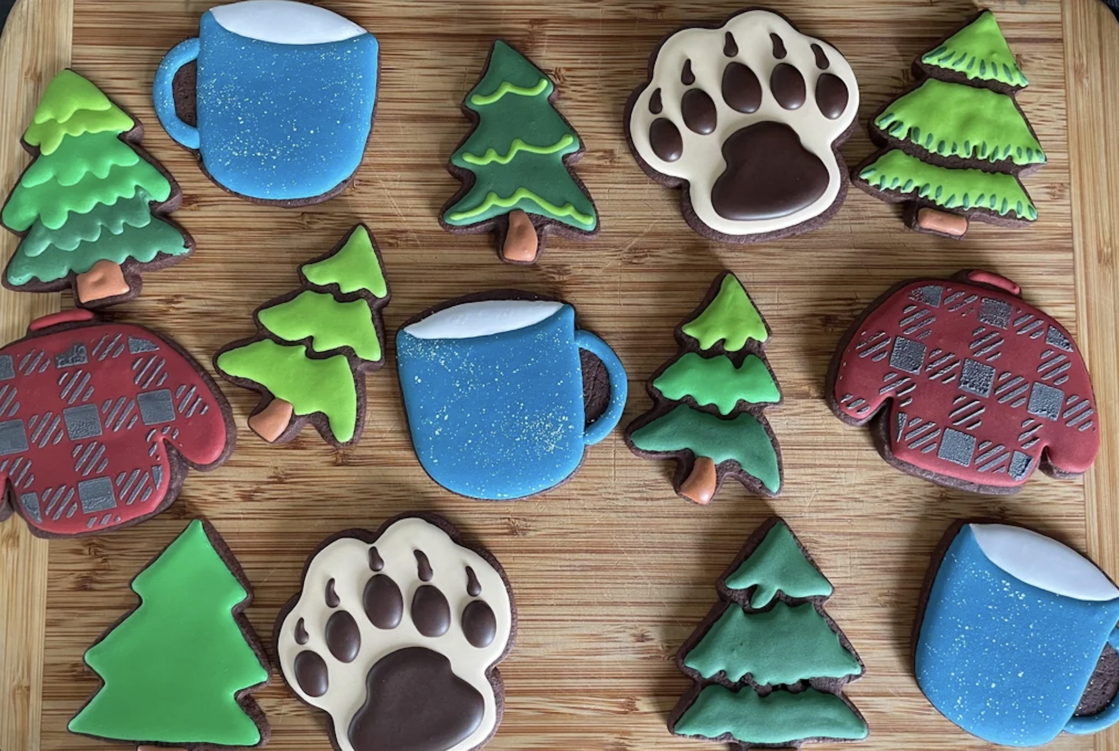 Iced cookies shaped like blue mugs, red mittens, green trees, and brown bear paw prints are arranged on a wooden surface.