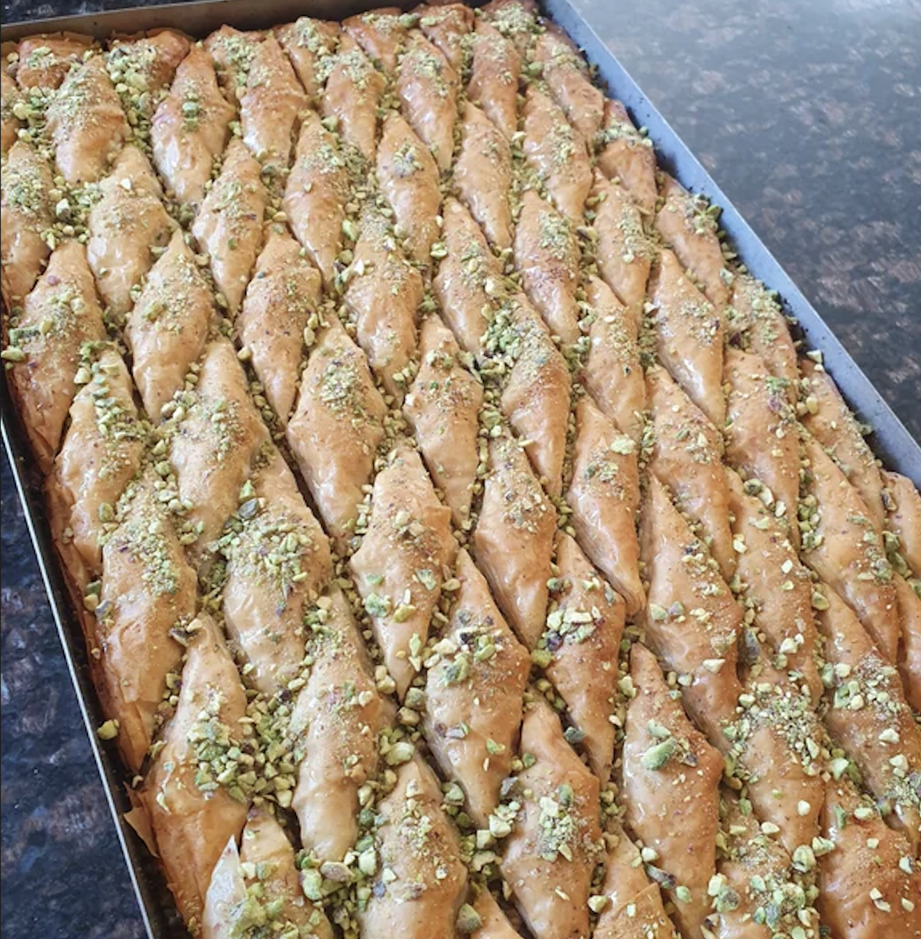 A tray filled with diamond-shaped pieces of baklava topped with chopped pistachios, arranged in neat rows on a dark countertop.