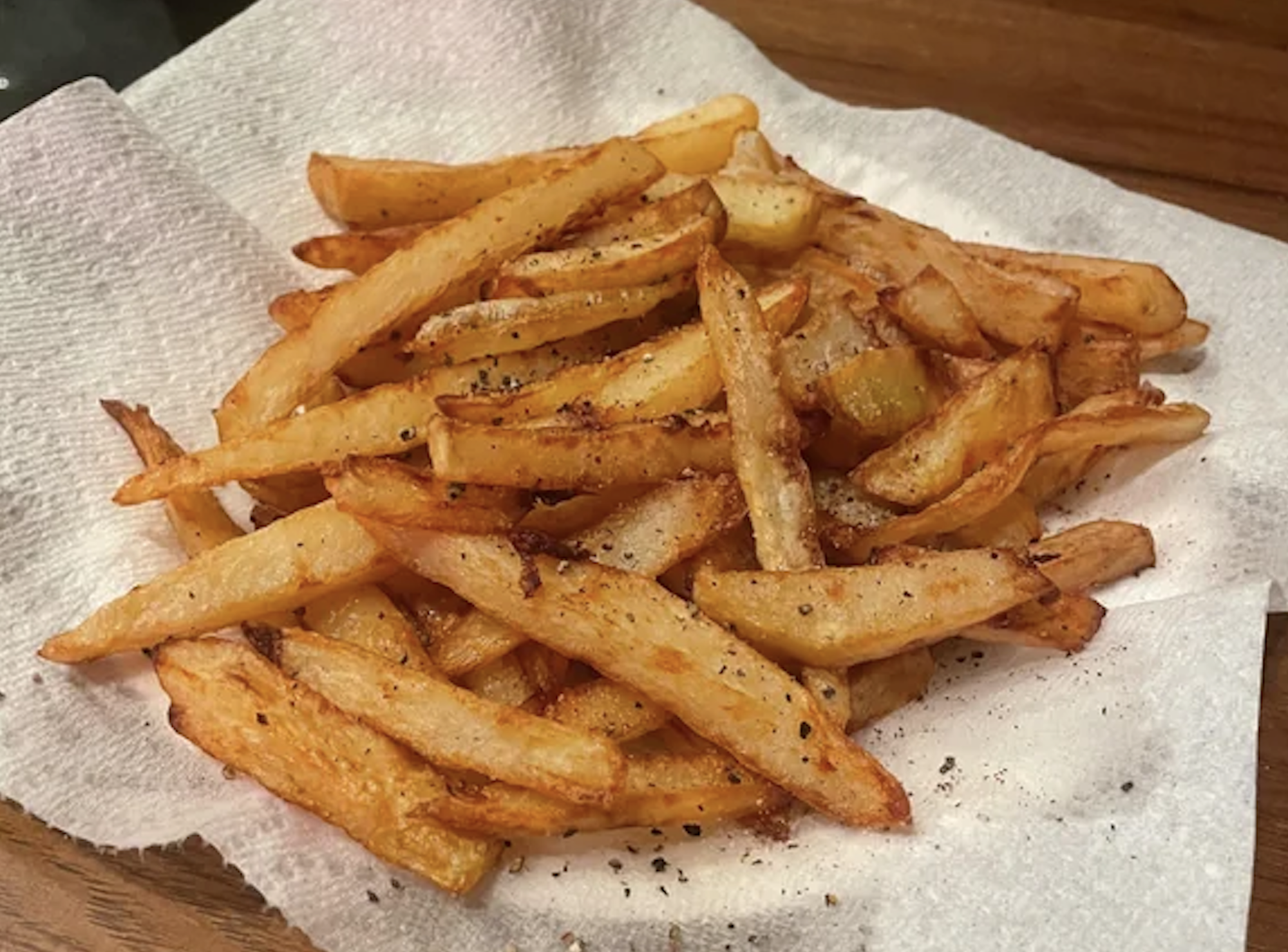 A pile of golden-brown French fries sprinkled with pepper rests on a white paper towel atop a wooden surface.