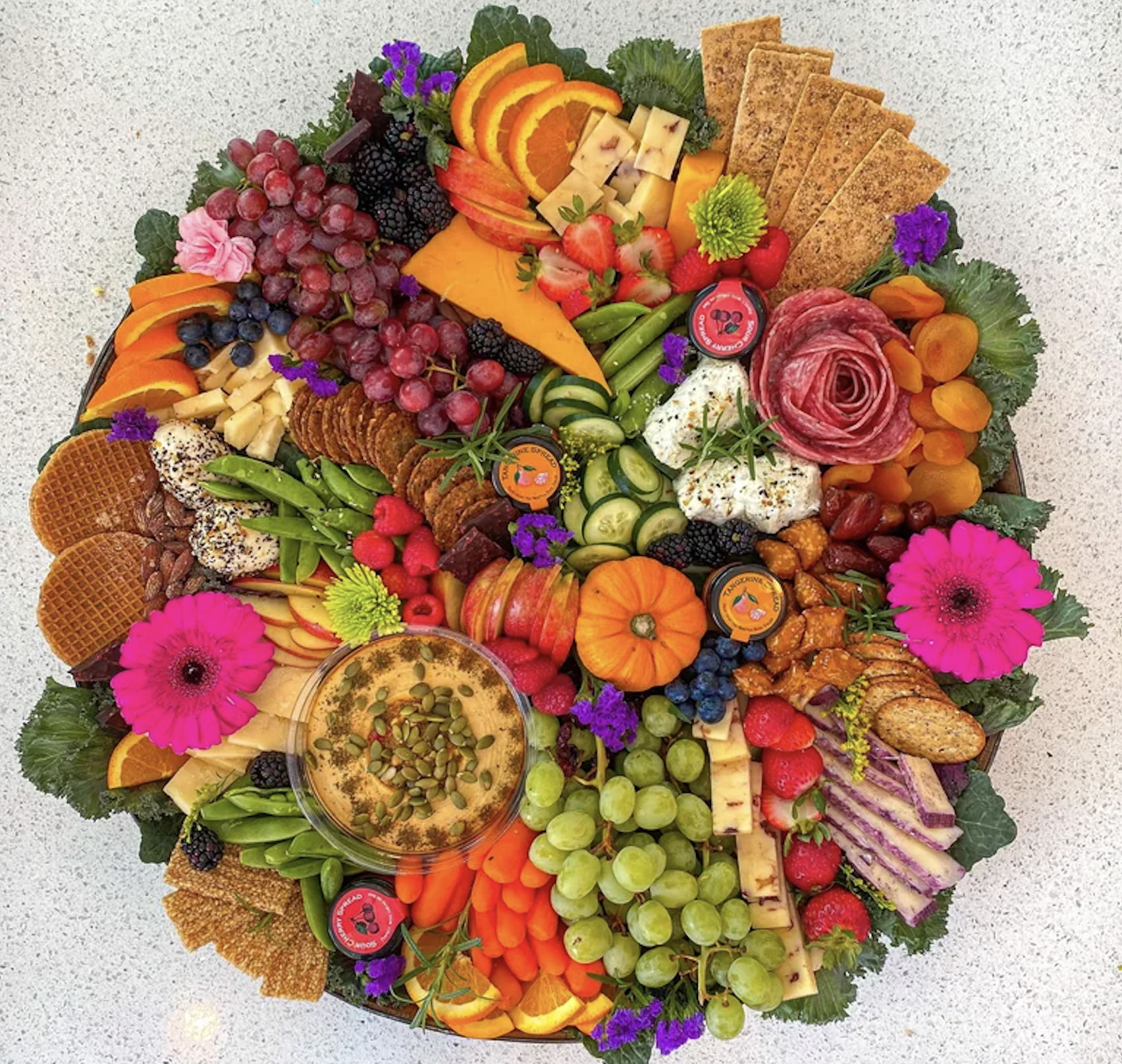 A colorful charcuterie board with assorted fruits, vegetables, cheeses, crackers, nuts, spreads, flowers, and mini pumpkins, arranged in a circular pattern on a white surface.