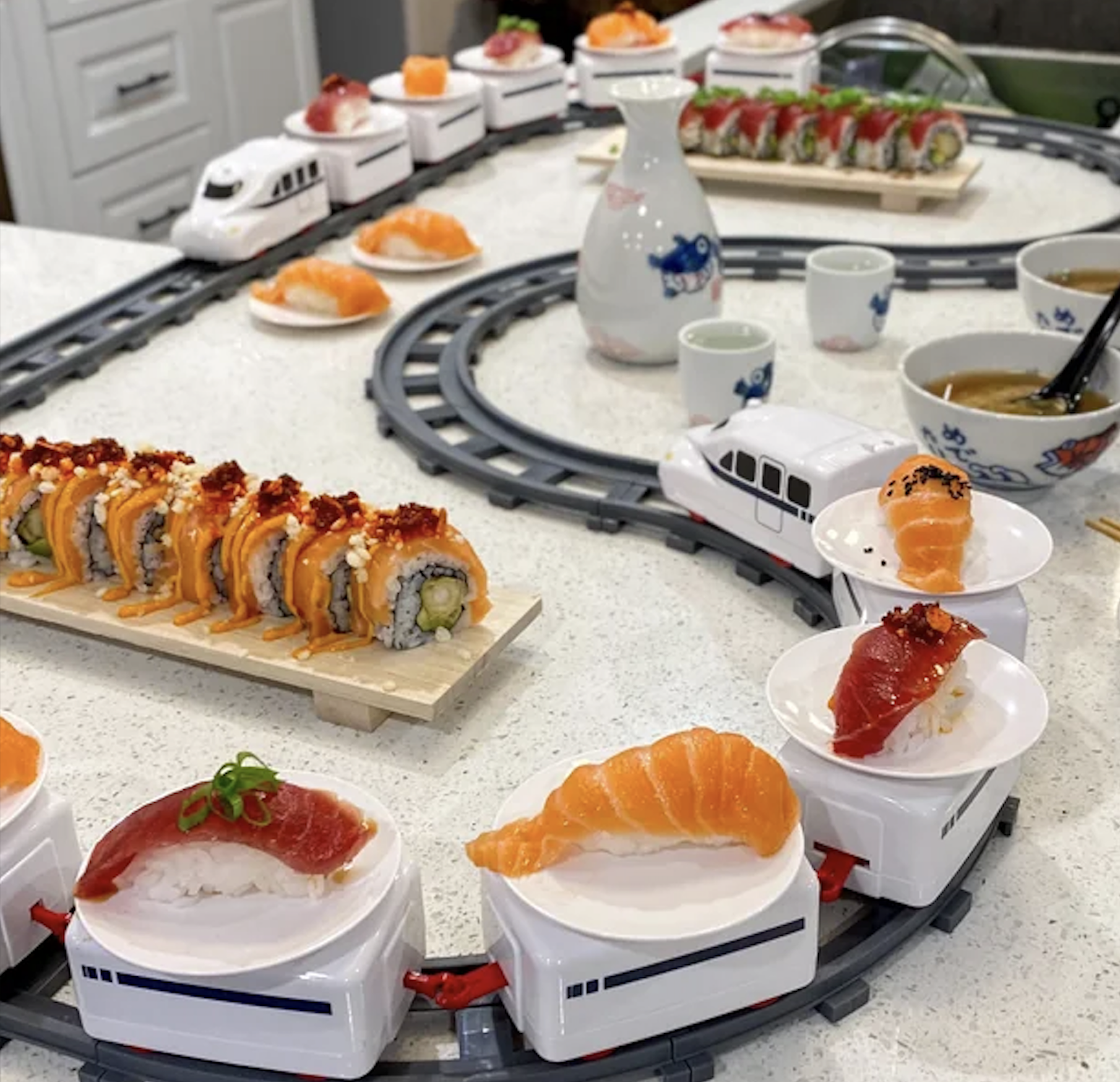 Toy trains deliver plates of sushi and nigiri around a table; sushi rolls, miso soup, and sake cups are also visible. The setup mimics a sushi train restaurant in a playful, home environment.