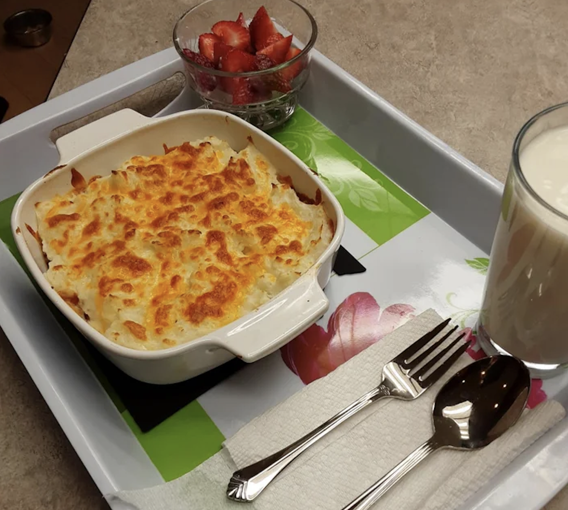A tray with a baked casserole, a glass of milk, a bowl of sliced strawberries, and a napkin with a fork and spoon placed beside the dishes.