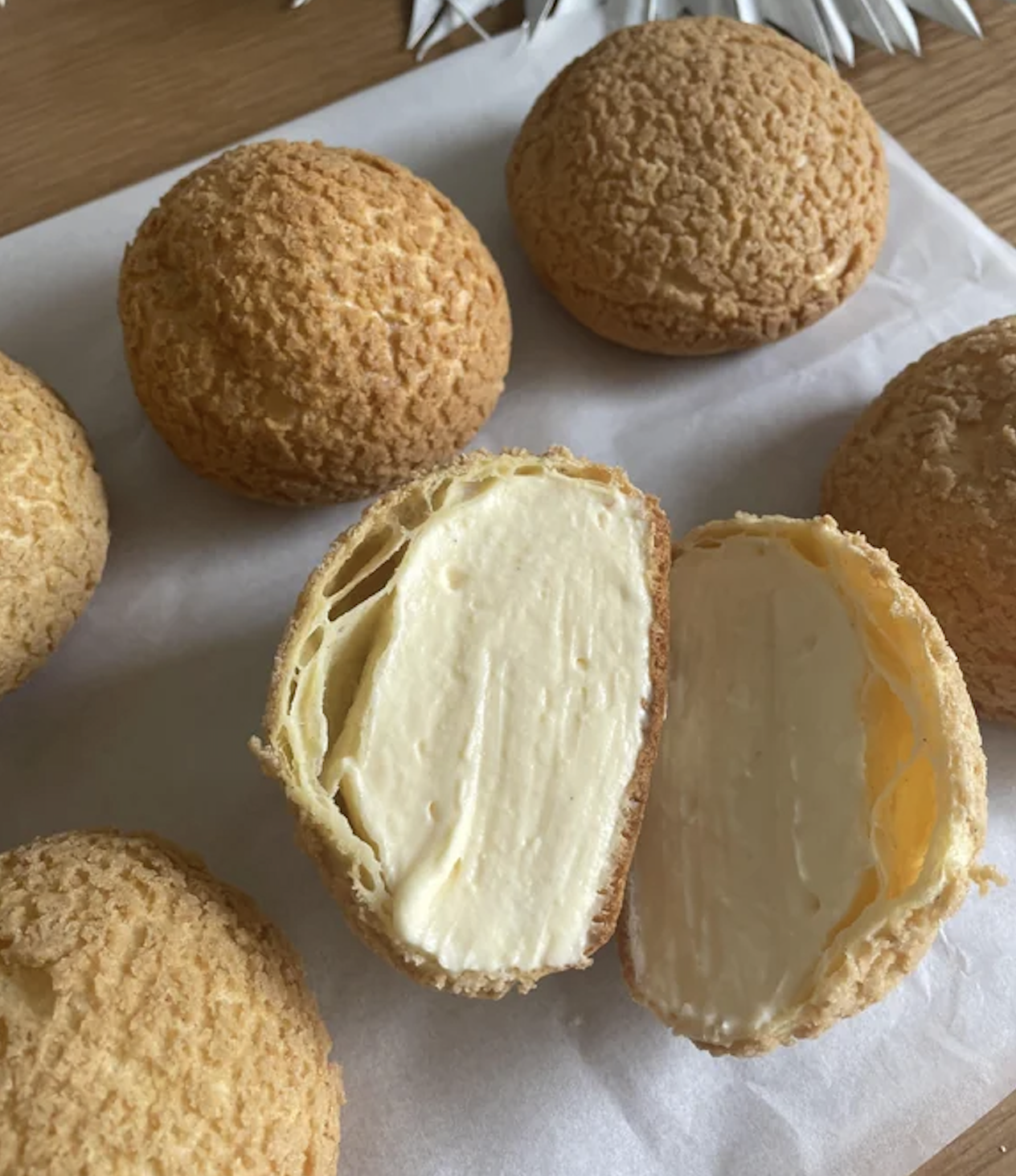 Six round cream puffs sit on parchment paper; one is cut open to reveal a thick filling of smooth vanilla cream inside the golden pastry shell.