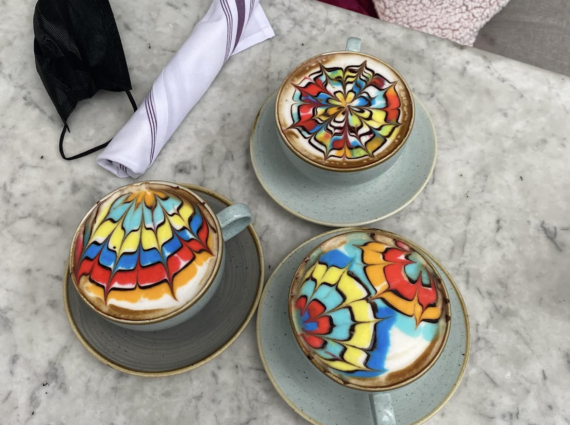 Three cups of coffee with colorful, intricate latte art sit on a marble table next to a rolled napkin and a black face mask.