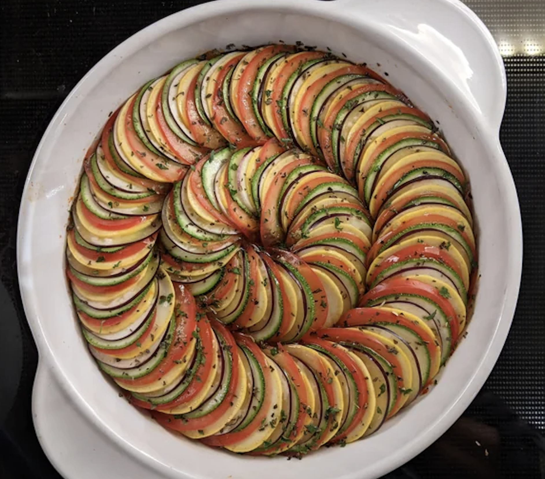 A round white baking dish filled with thinly sliced zucchini, yellow squash, and tomato, arranged in a tight spiral pattern and topped with herbs.