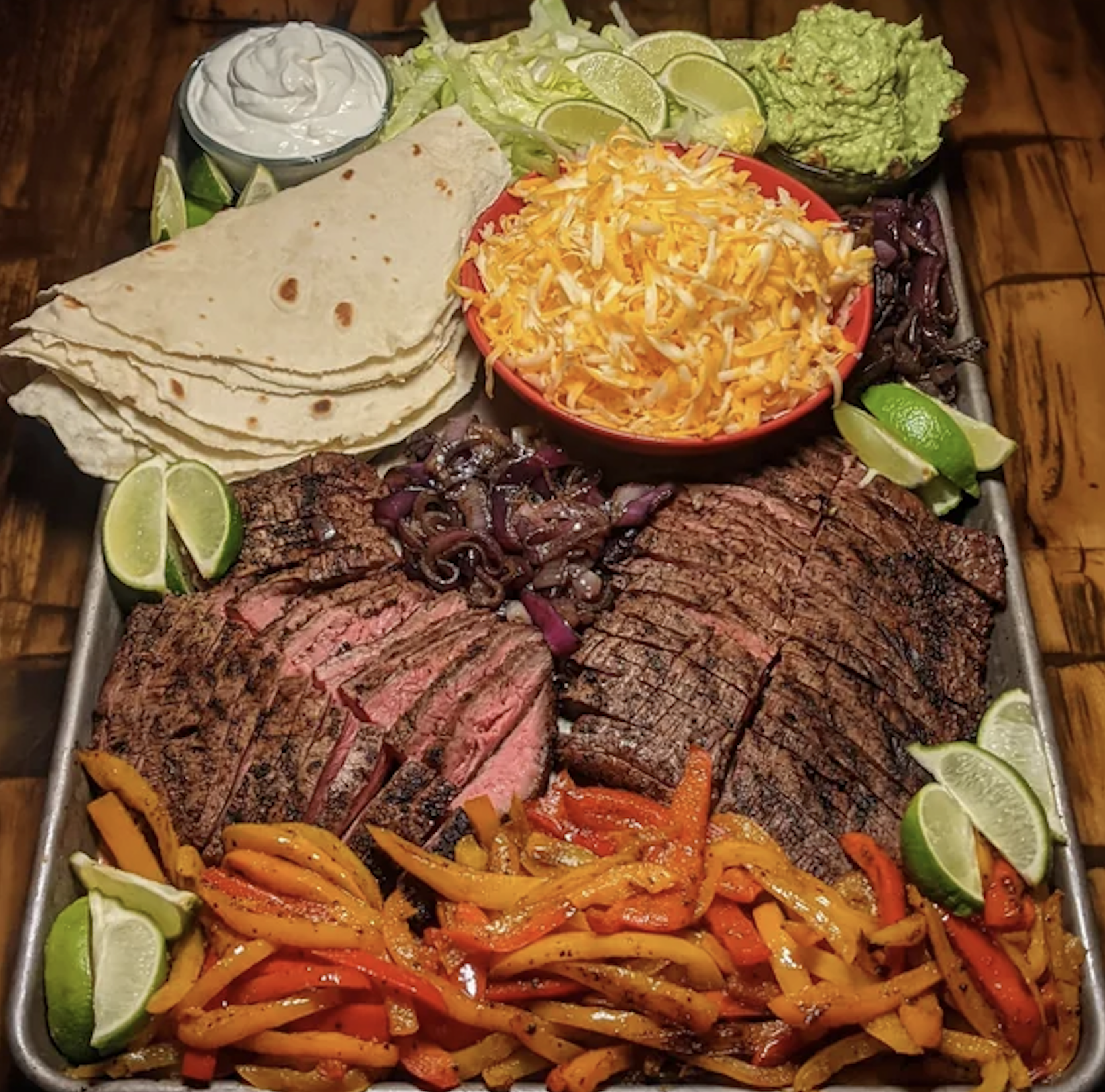 A tray with sliced grilled steak, sautéed peppers and onions, shredded cheese, lettuce, lime wedges, tortillas, sour cream, and guacamole, arranged neatly on a wooden background.