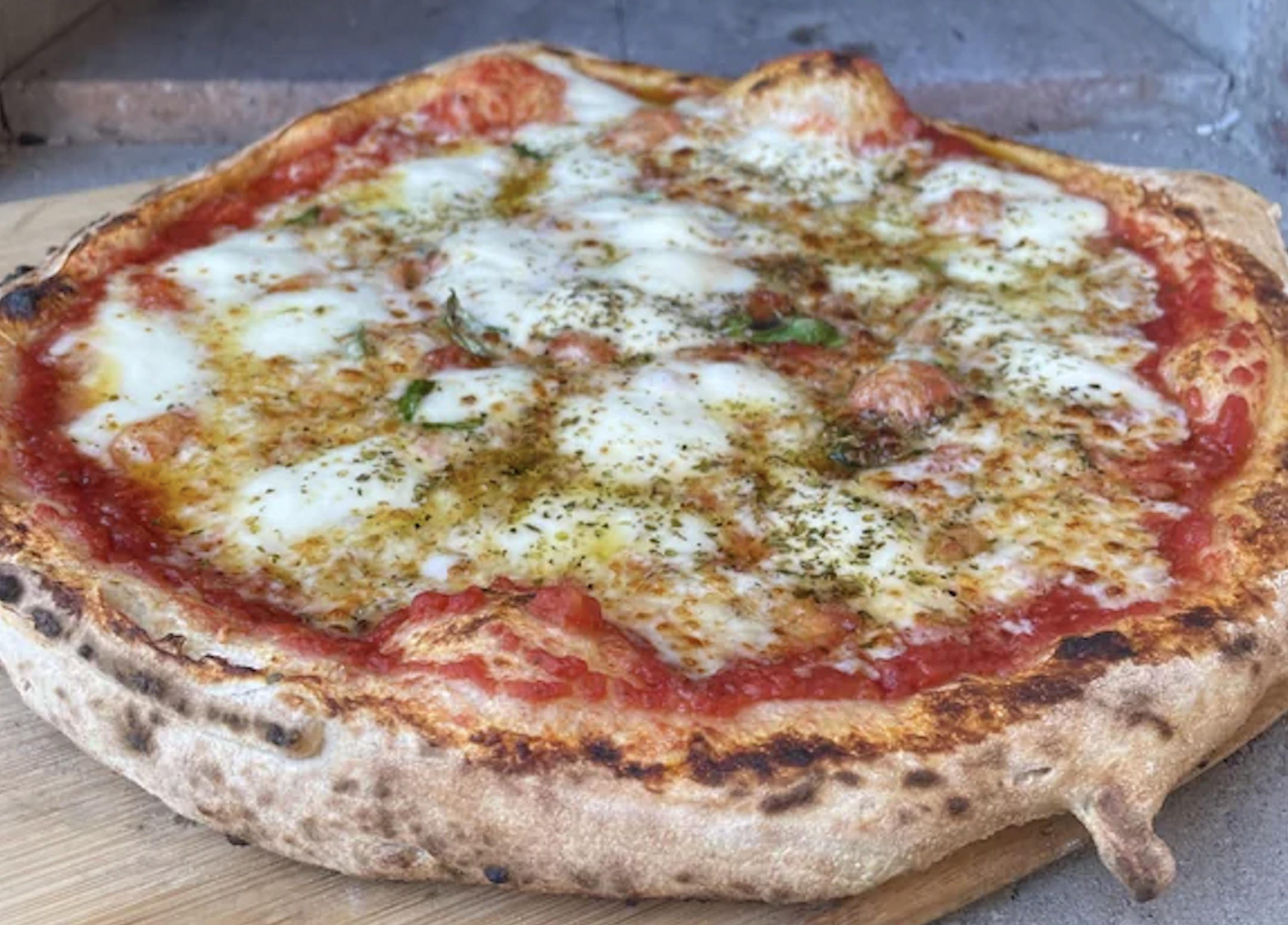 A freshly baked pizza with melted cheese, tomato sauce, herbs, and a golden, slightly charred crust sits on a wooden board.