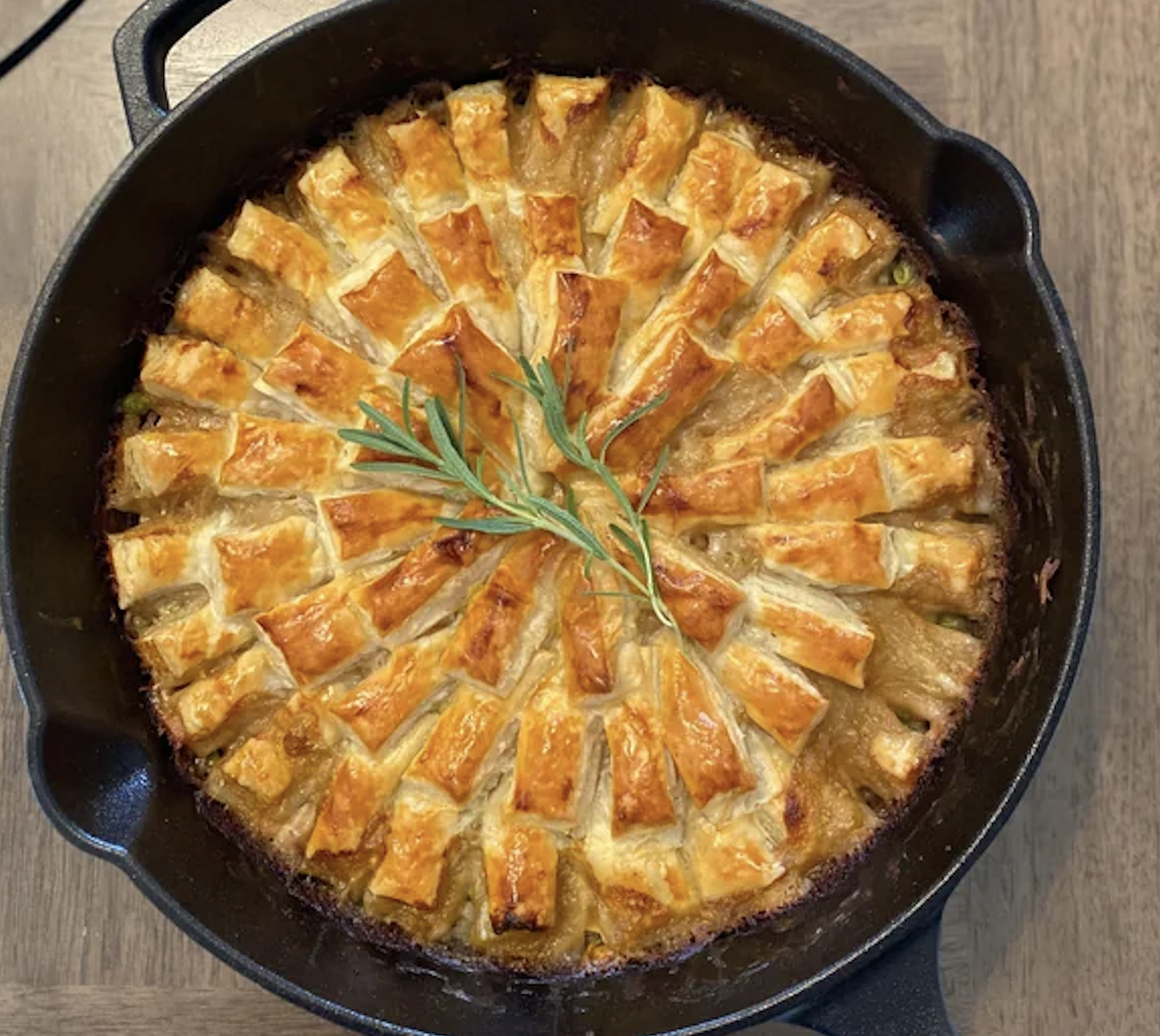 A golden, lattice-topped pie baked in a cast iron skillet, garnished with a sprig of fresh rosemary in the center, sits on a wooden table.
