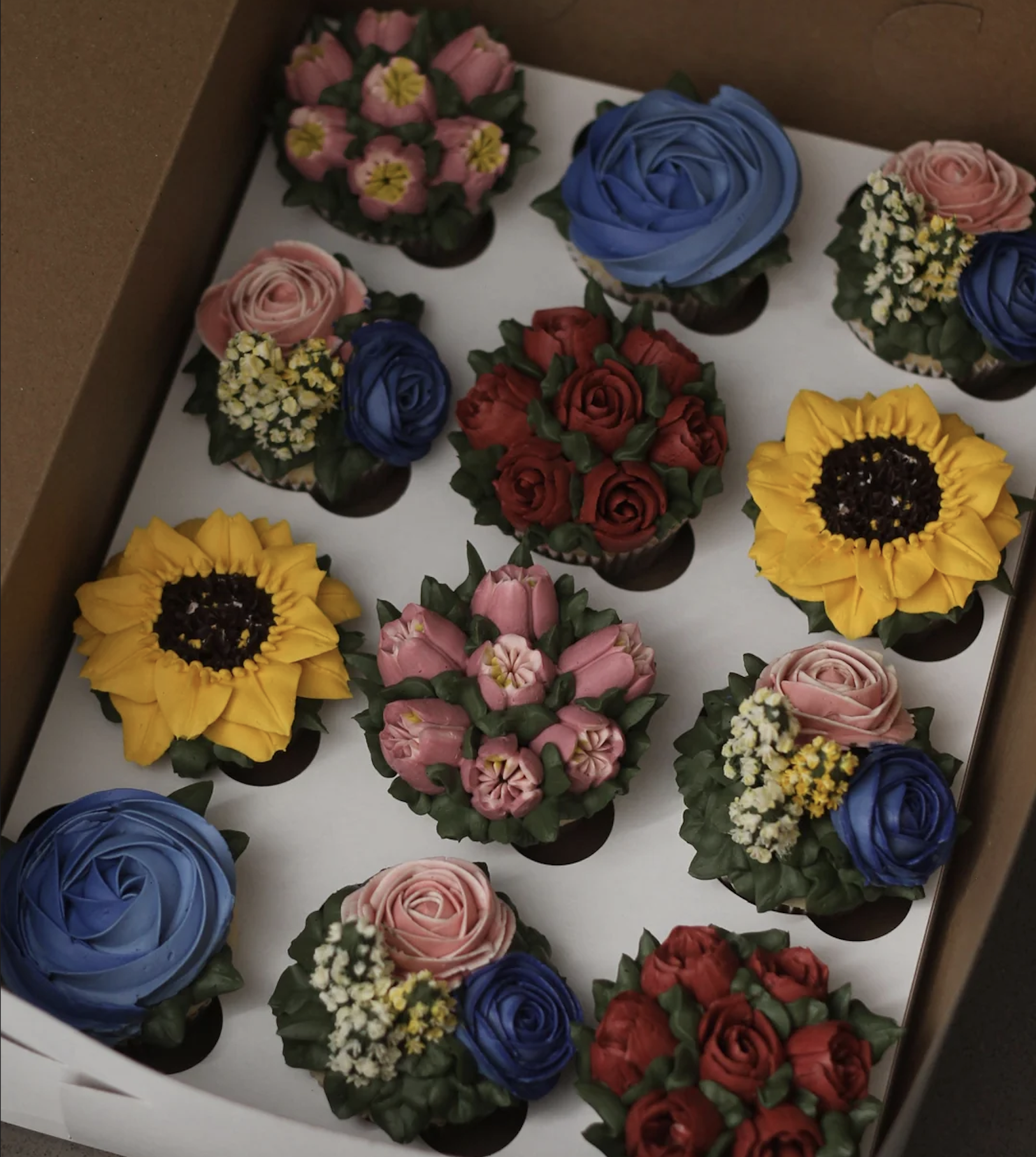 A box of twelve cupcakes decorated with colorful buttercream flowers, including sunflowers, blue roses, red roses, pink blossoms, and small white and yellow flowers.