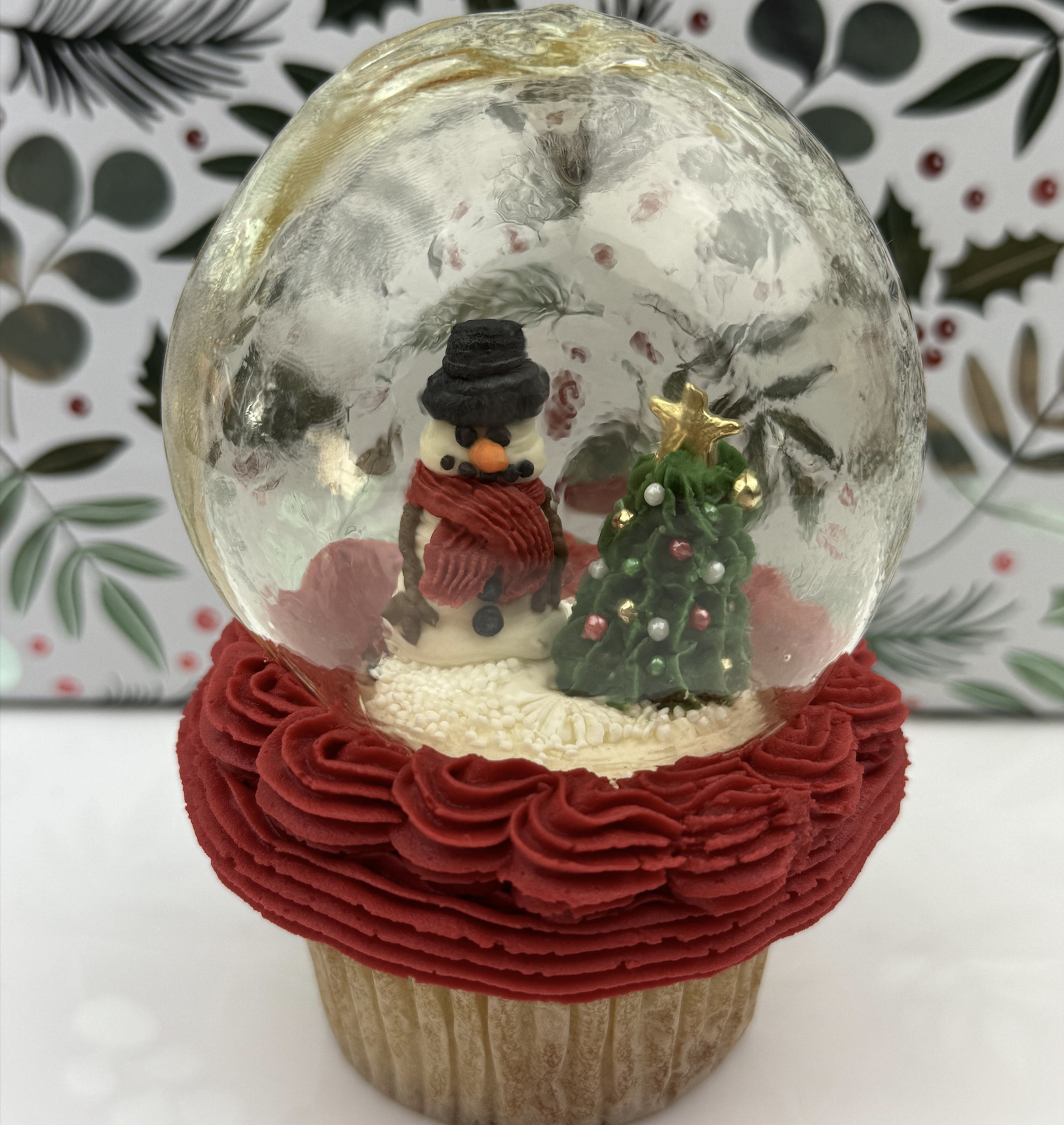 A cupcake topped with red frosting, a clear sugar dome, a small snowman wearing a hat and scarf, and a decorated Christmas tree inside. Holiday-themed background with green leaves and red berries.