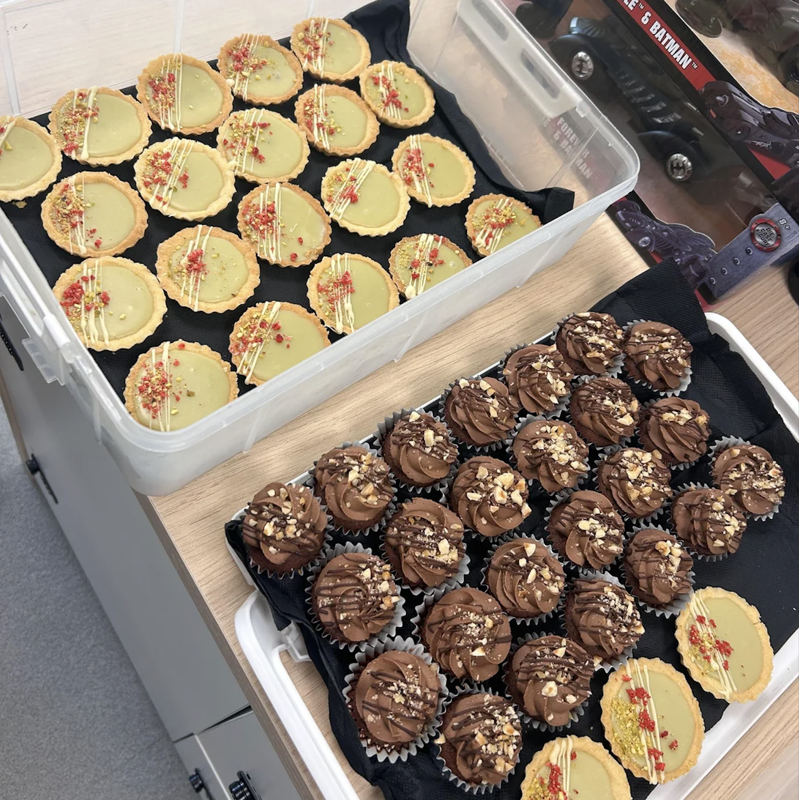 Two trays of cupcakes: one with yellow icing and red sprinkles, the other with chocolate icing and nut pieces. Both trays are placed on a wooden surface.