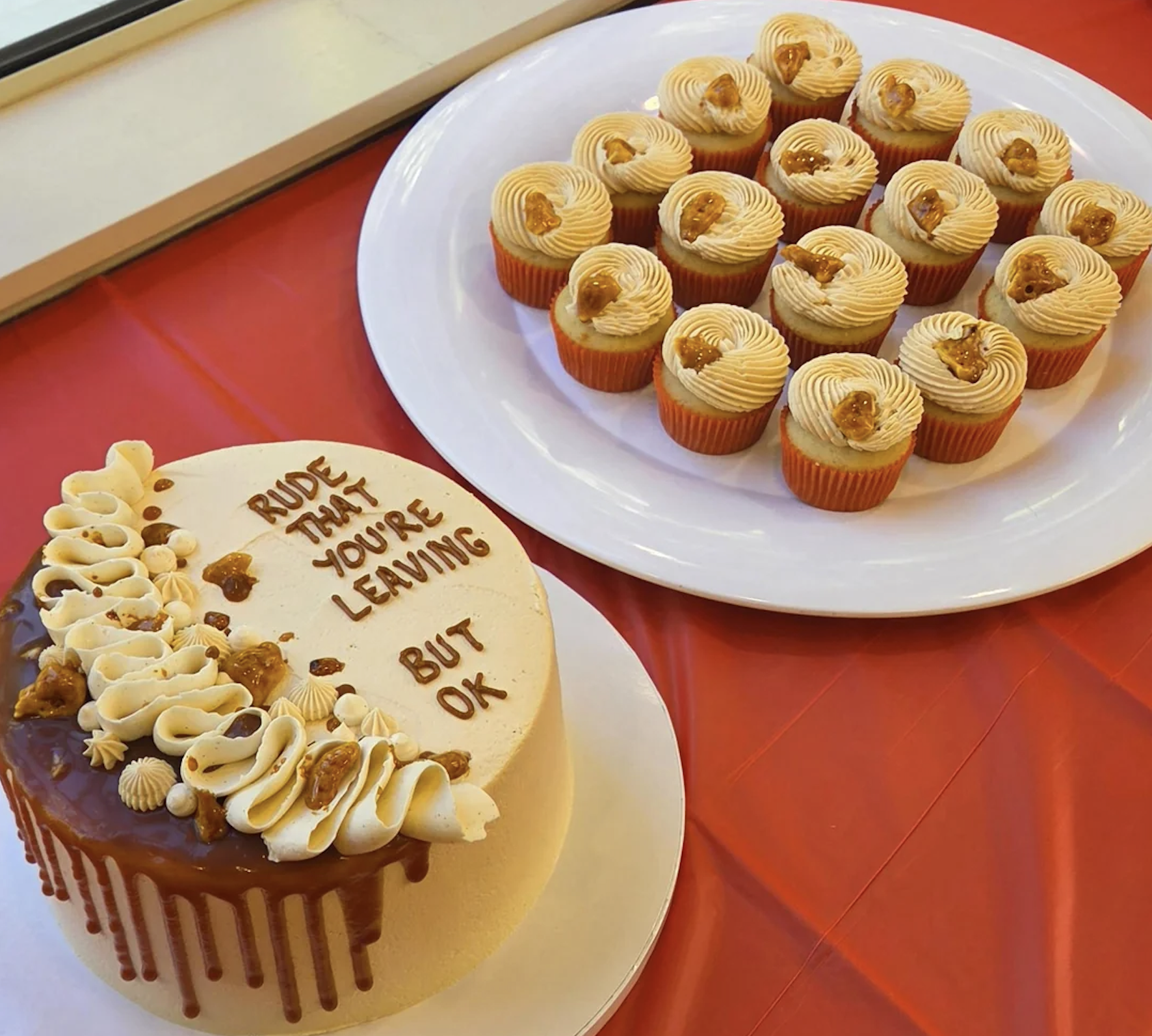A round cake with “Rude that you’re leaving but OK” written on top is next to a plate of cupcakes with swirled frosting, all on a red tablecloth.