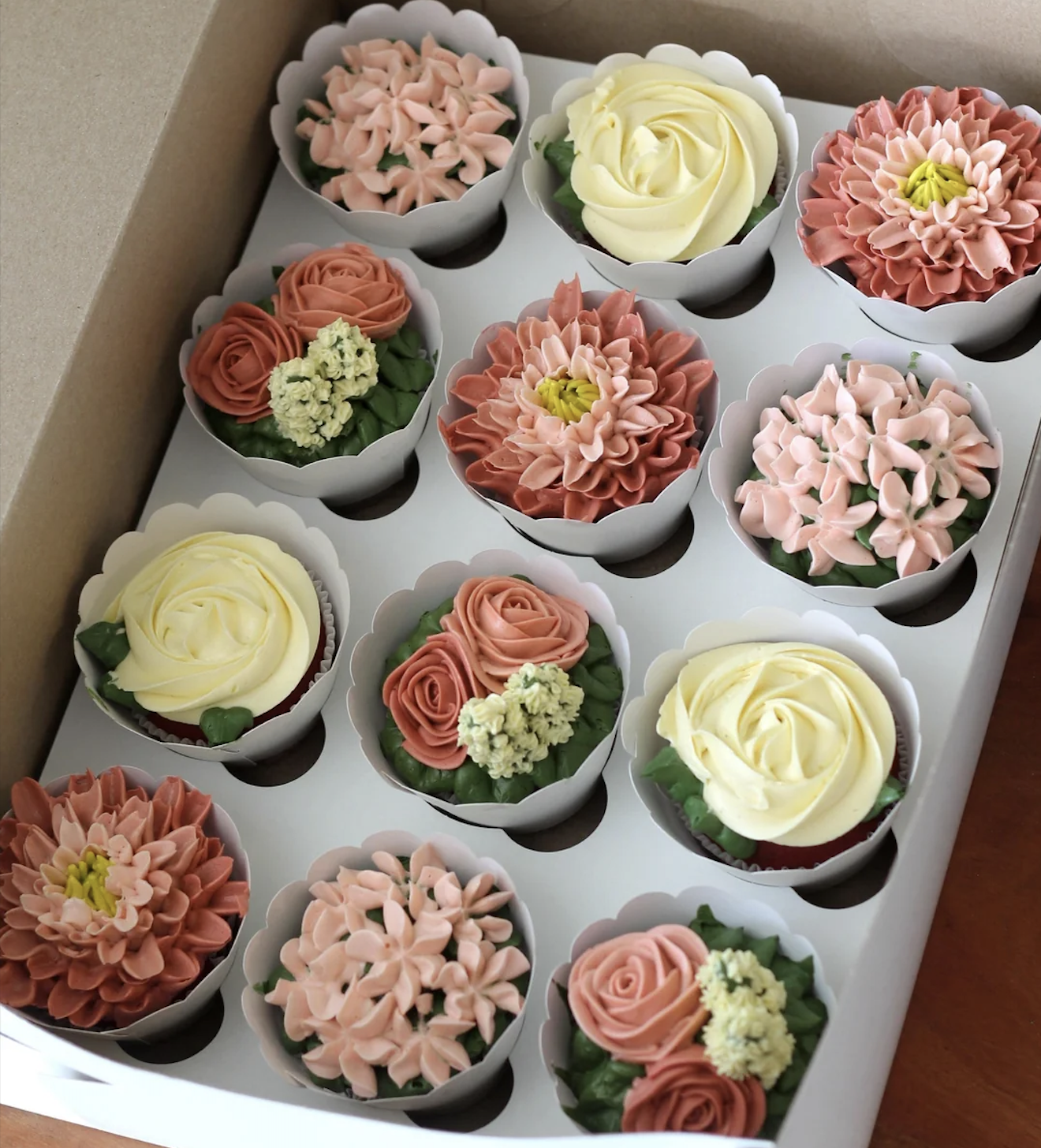 A box of twelve cupcakes decorated with intricate buttercream flowers in soft shades of pink, peach, cream, and green, arranged neatly in three rows.
