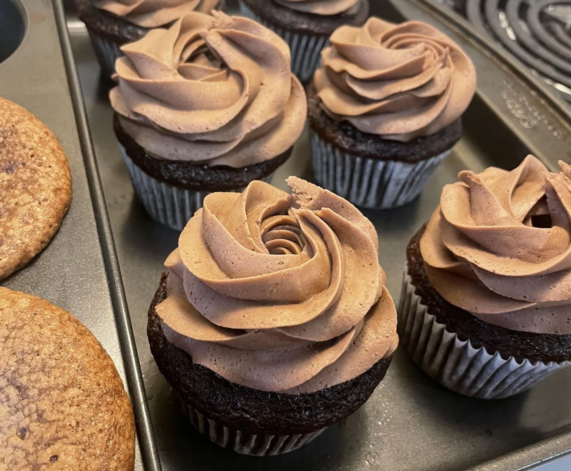 Chocolate cupcakes with swirled chocolate frosting sit in a muffin tin next to some cookies. The cupcakes have paper liners and the frosting is piped in a rose pattern.
