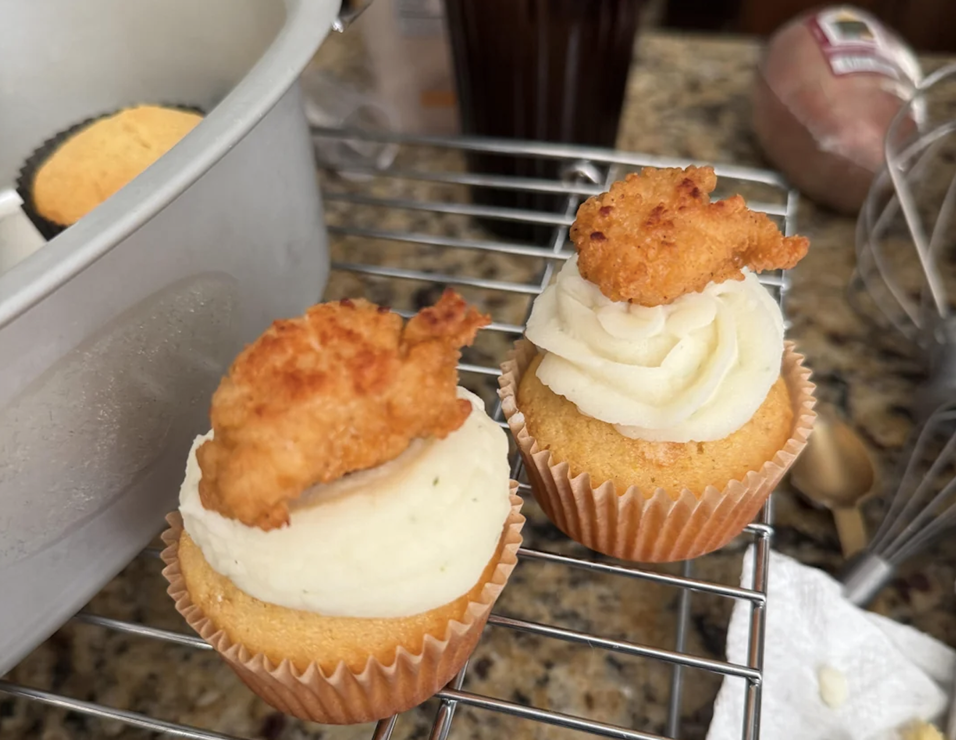 Two cupcakes with white frosting, each topped with a piece of fried chicken, sit on a cooling rack in a kitchen setting with baking tools in the background.
