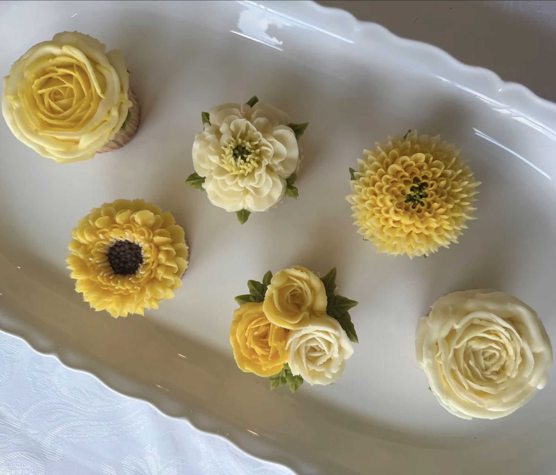 Six cupcakes decorated with yellow and white buttercream flowers, displayed on a white rectangular tray with scalloped edges. The flowers resemble roses, dahlias, and sunflowers.