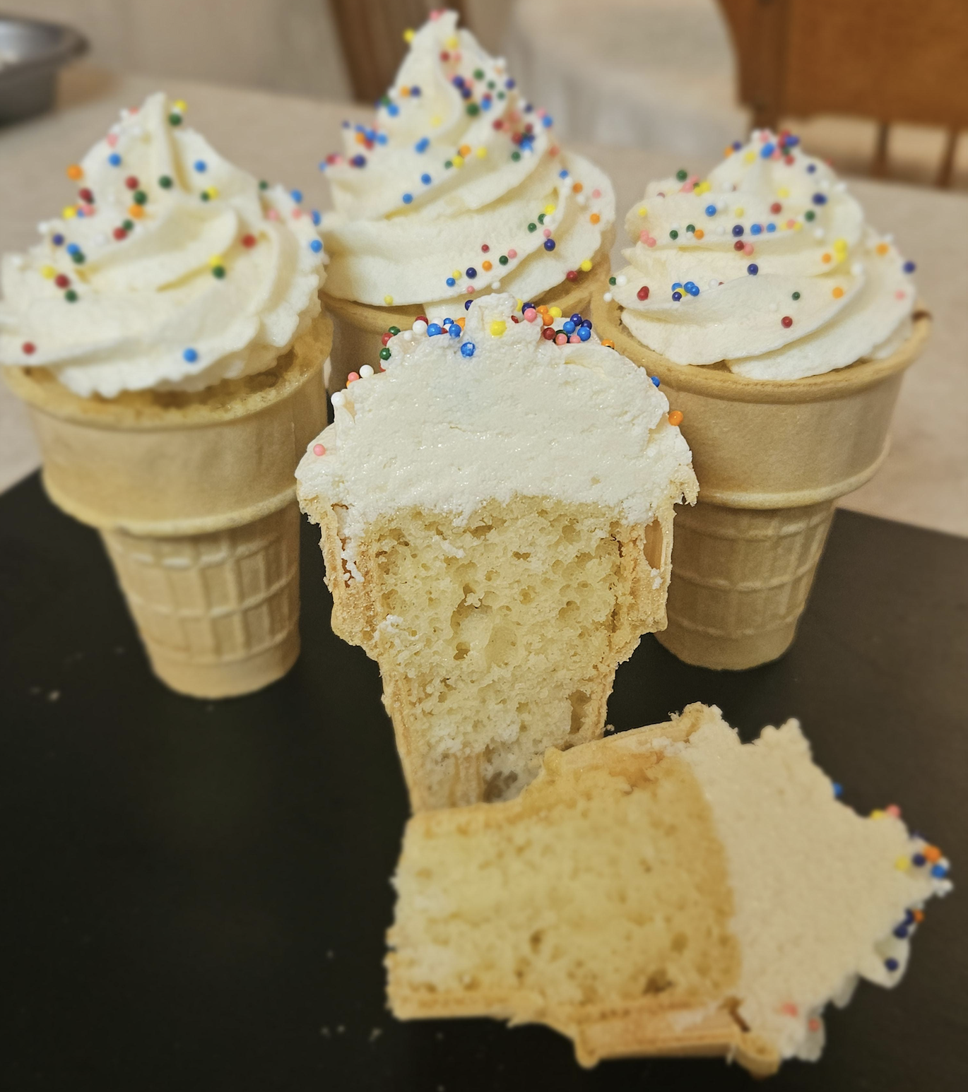 Four ice cream cone-shaped cupcakes with white swirled frosting and colorful sprinkles; one is cut in half to show the cake inside.