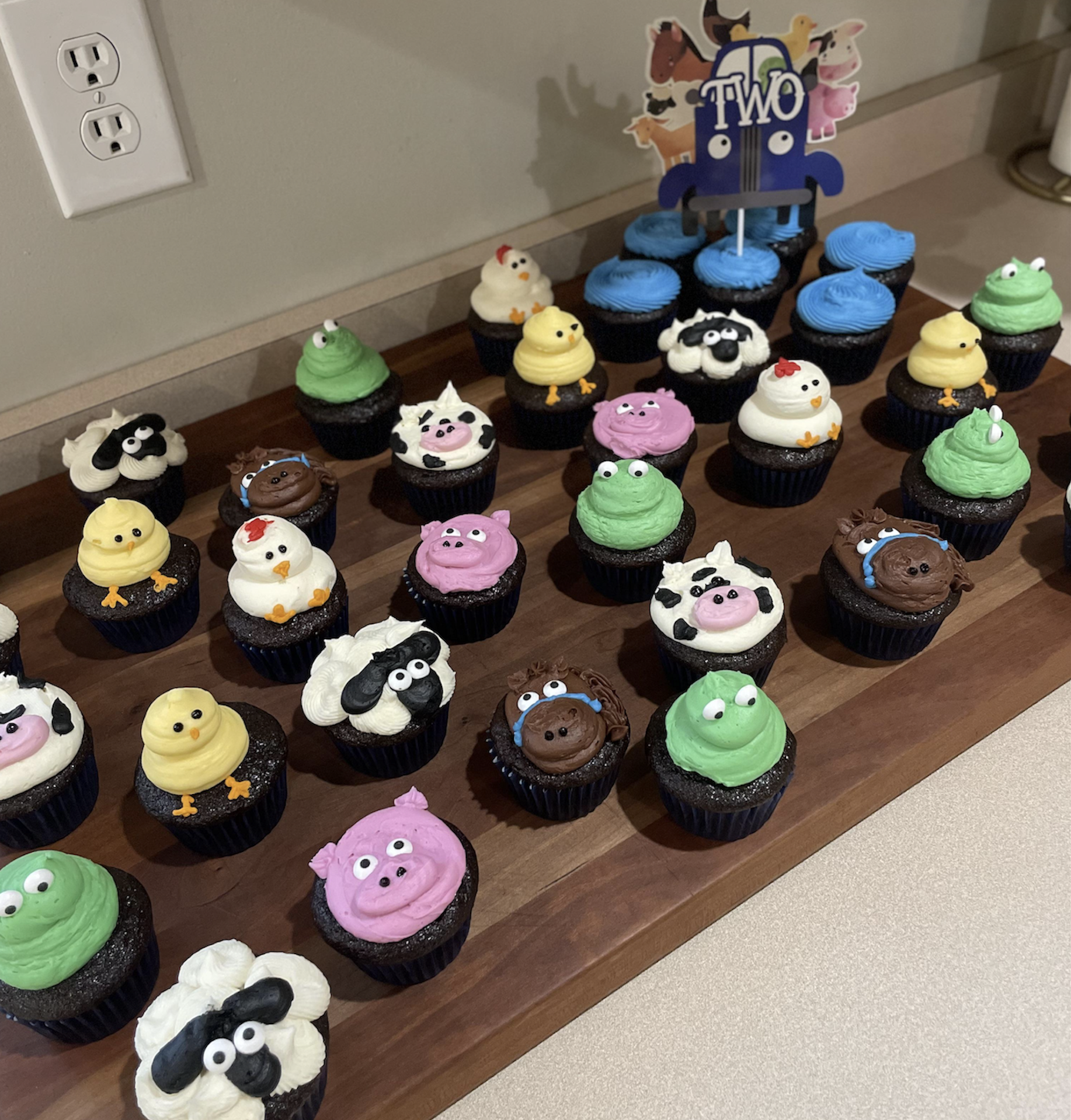 A tray of cupcakes decorated as farm animals, including pigs, cows, chicks, and sheep, with a “TWO” sign on blue cupcakes, indicating a child’s second birthday celebration.