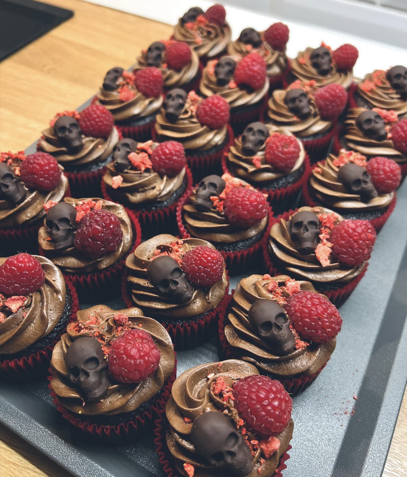 Rows of chocolate cupcakes topped with chocolate skulls, fresh raspberries, and red crumble, displayed on a dark tray on a wooden surface.