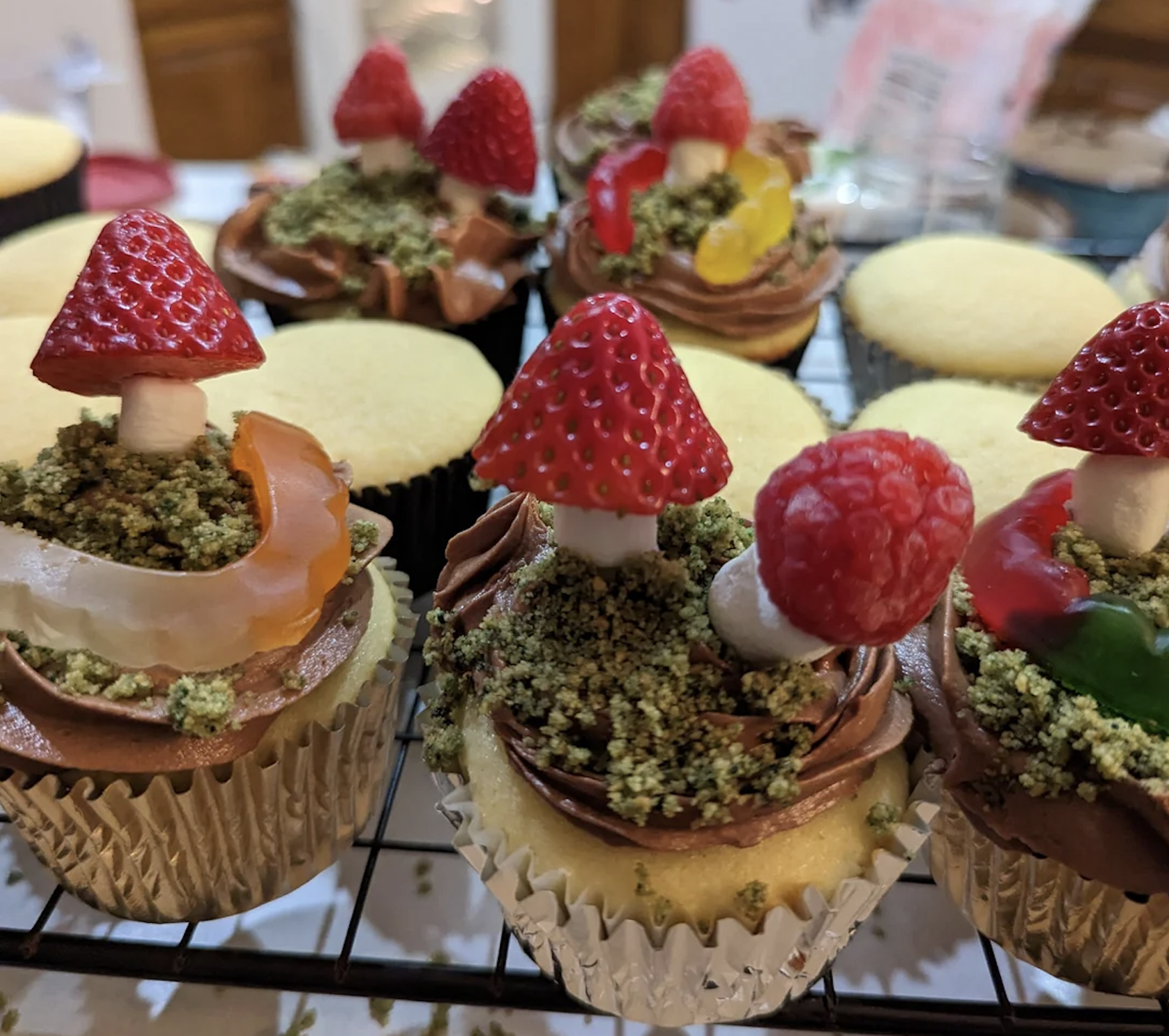 Vanilla cupcakes decorated with chocolate frosting, green crumbs, and fruit shaped like mushrooms; strawberry and raspberry tops on marshmallow stems, with gummy candy accents. Other plain cupcakes are visible in the background.