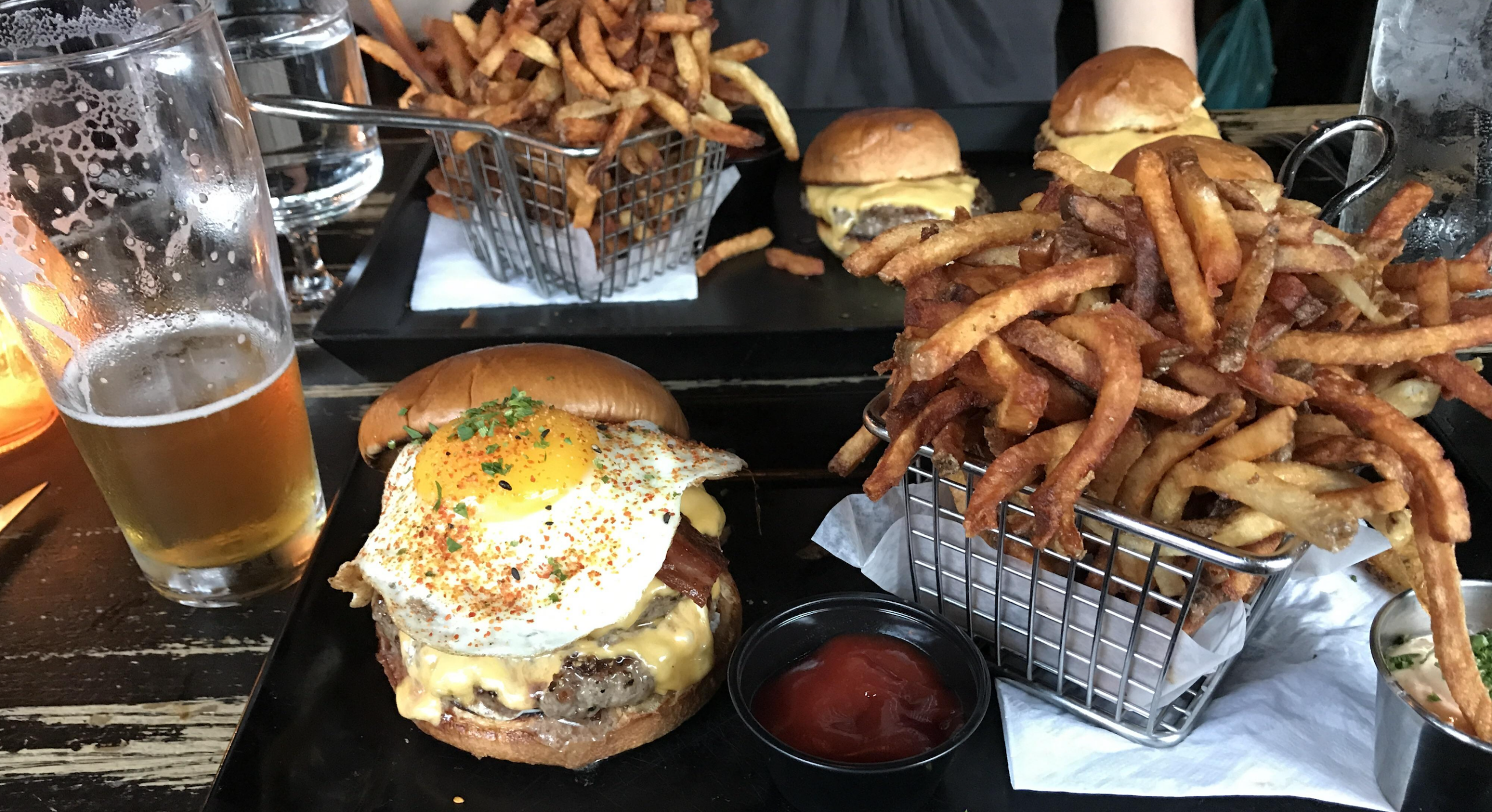 A table with burgers, fries in metal baskets, a glass of beer, and a burger topped with a fried egg, cheese, and bacon, served with a cup of ketchup.