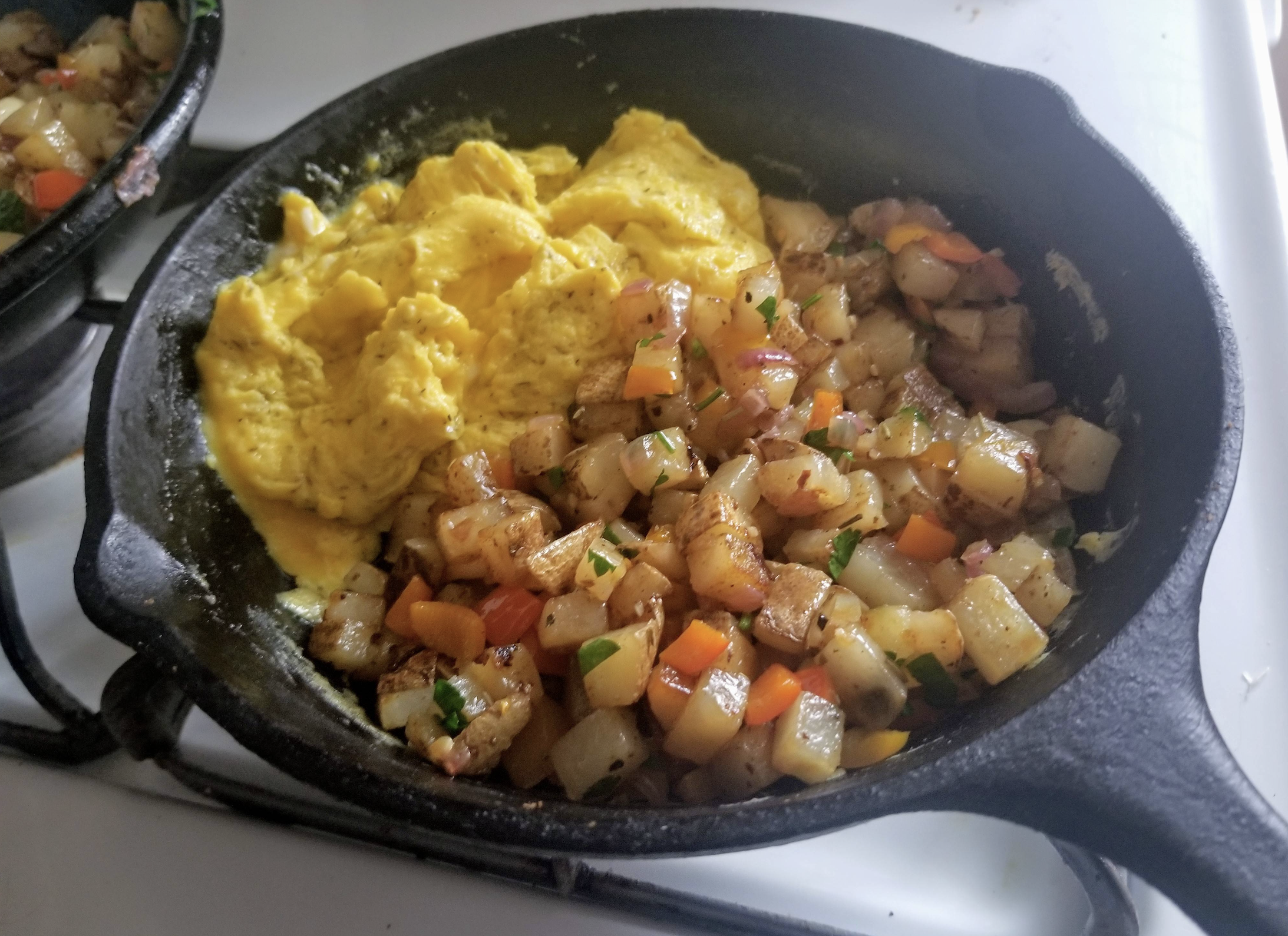 Scrambled eggs and diced potatoes with peppers and onions cook together in a black cast iron skillet on a stovetop.