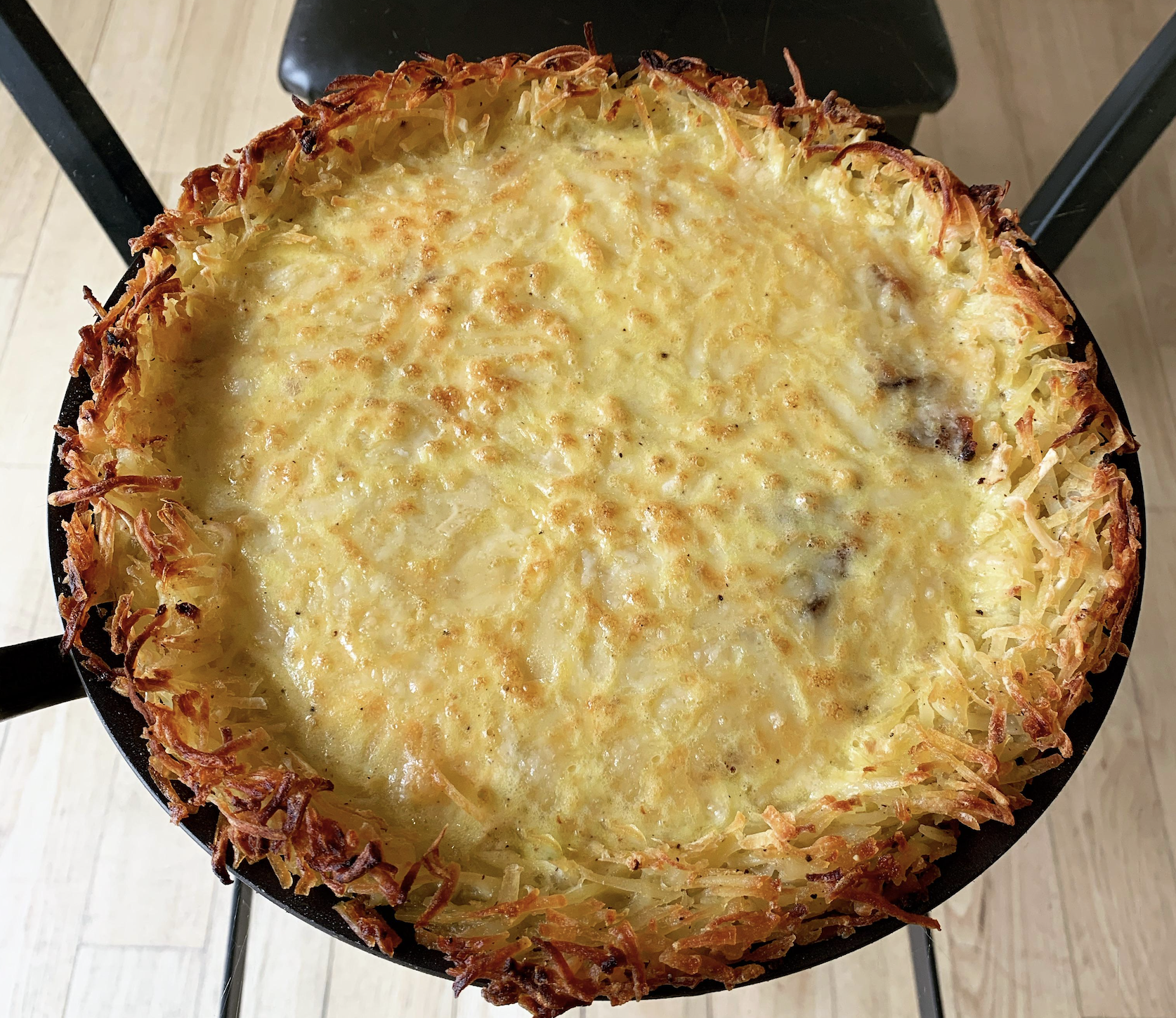 A baked dish in a round pan with a golden, crispy edge and a cheesy, lightly browned top, sitting on a black chair and light wooden floor.