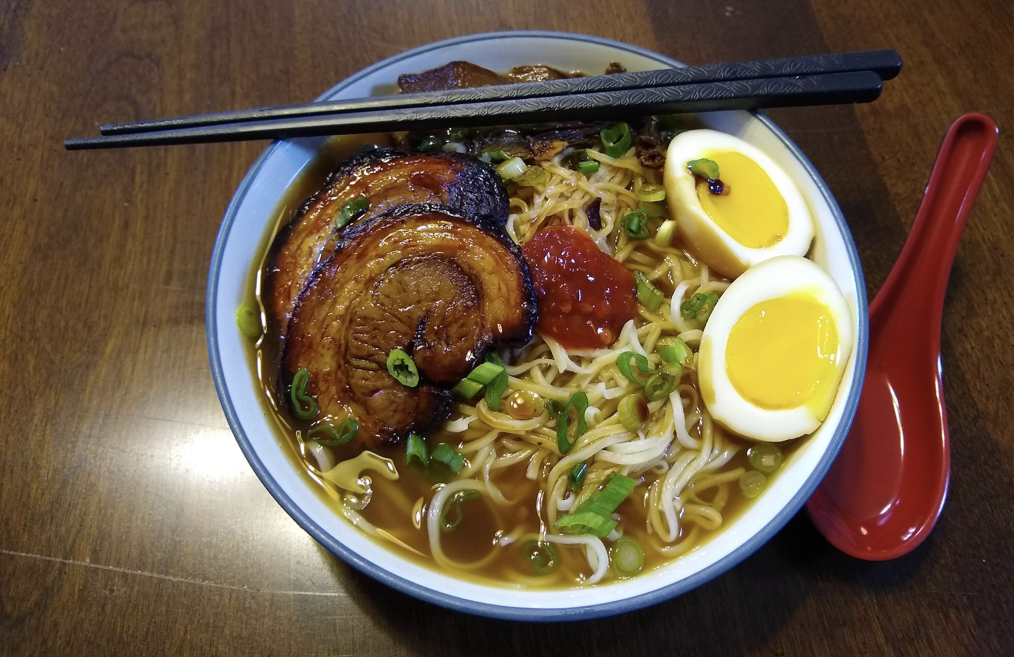 A bowl of ramen with sliced pork, halved soft-boiled eggs, green onions, noodles, broth, and a dollop of chili paste, with chopsticks placed across the bowl and a red soup spoon beside it.