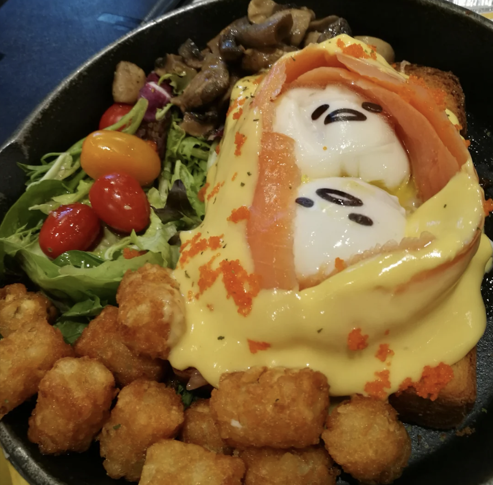 A skillet breakfast with tater tots, salad, tomatoes, mushrooms, and toast topped with two eggs decorated as cartoon faces, wrapped in smoked salmon and drizzled with yellow sauce.