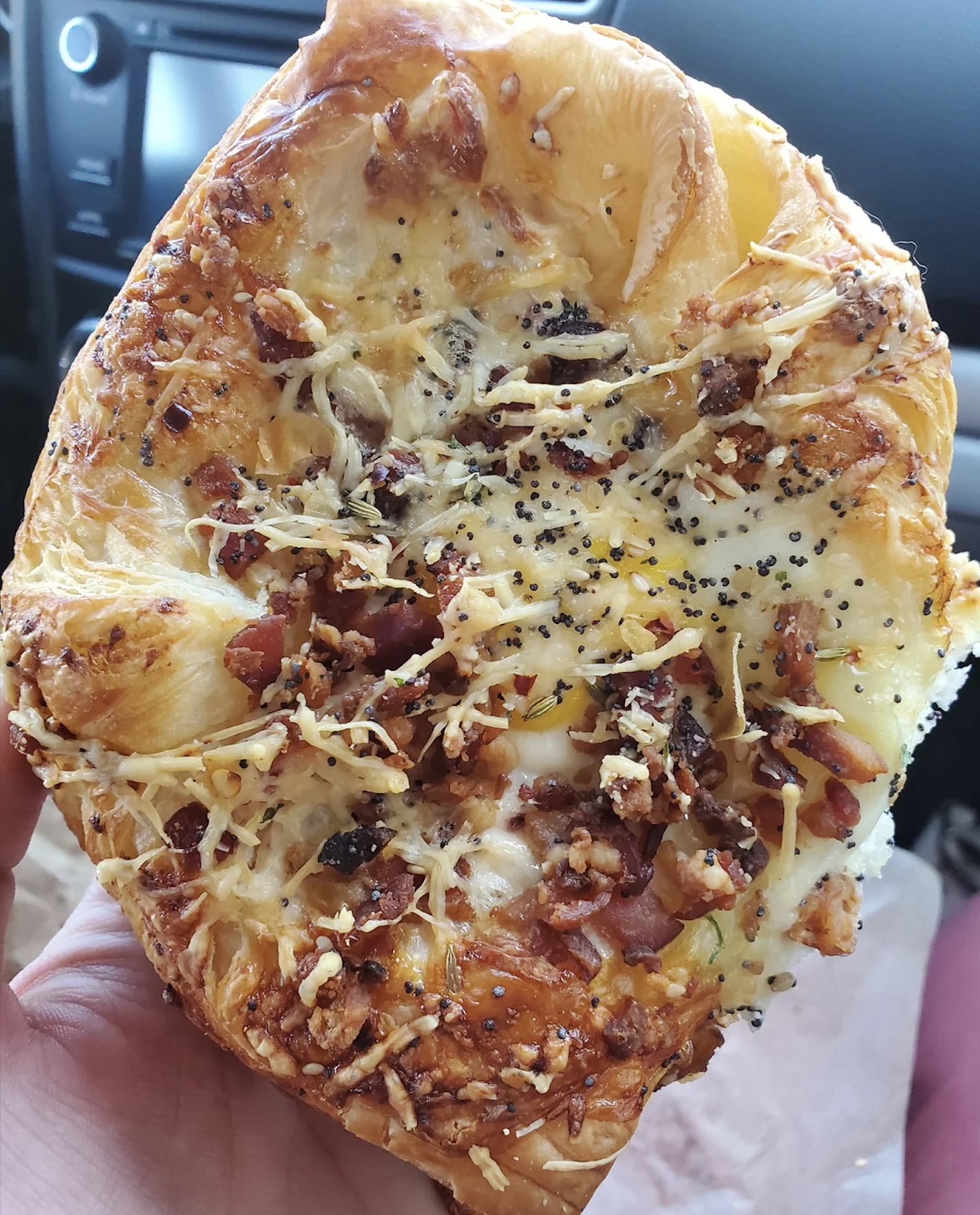 A hand holds a cheesy, savory pastry topped with shredded cheese, bacon bits, poppy seeds, and herbs, with a car dashboard visible in the background.