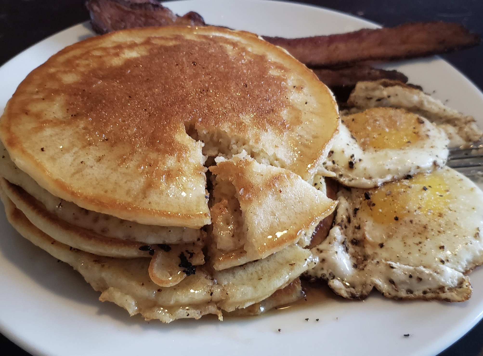 A plate with a stack of pancakes, a piece cut out, two sunny-side-up eggs, and strips of crispy bacon. Syrup is drizzled over the pancakes.