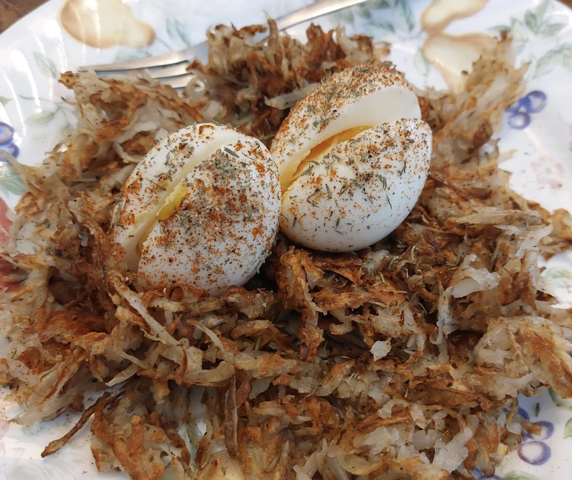 Two boiled eggs sprinkled with herbs and paprika sit on a bed of crispy hash browns on a floral-patterned plate, with a fork in the background.