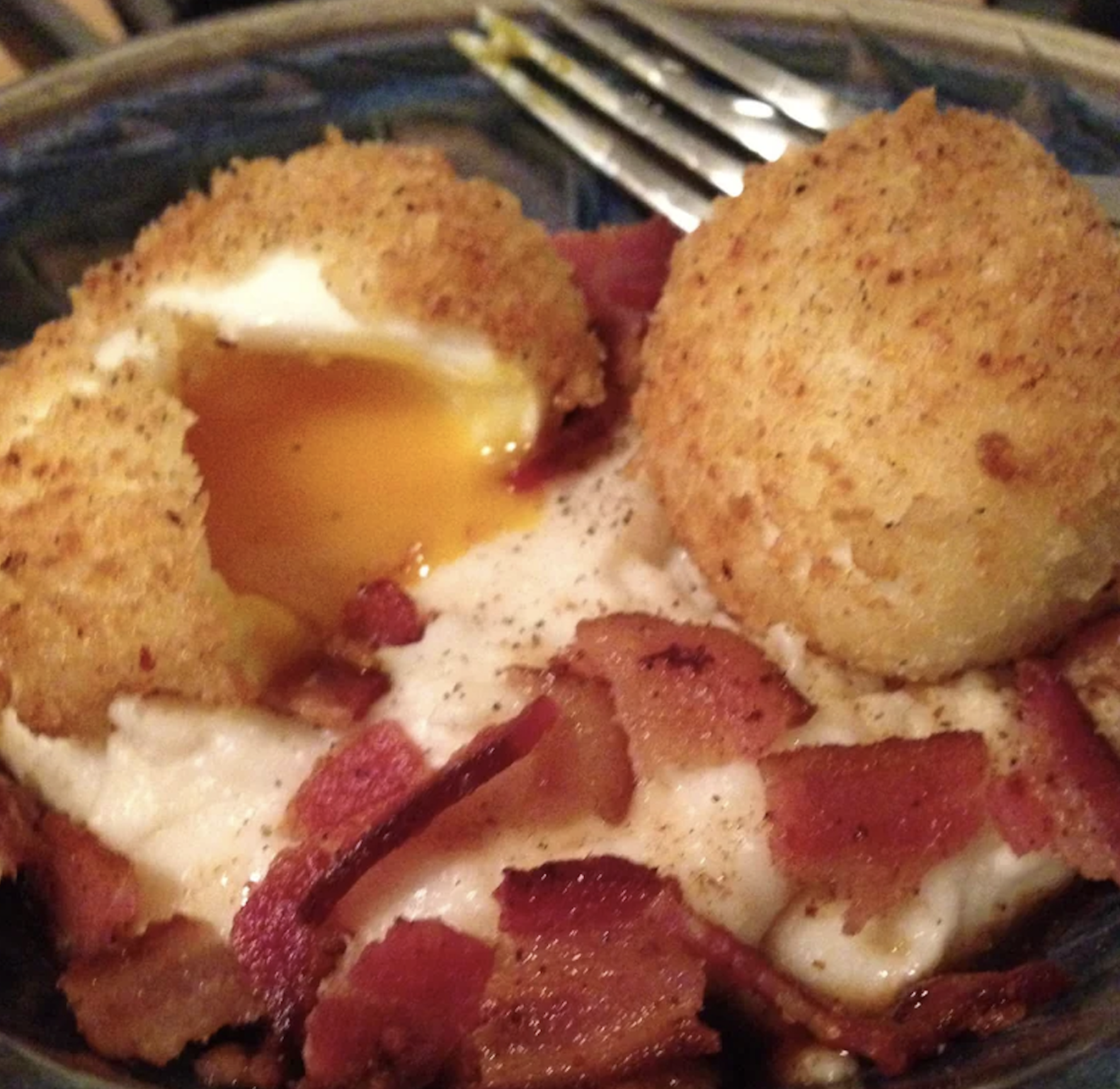 Two breaded and fried eggs, one with a runny yolk, sit on a plate with crispy bacon pieces. A fork rests in the background.