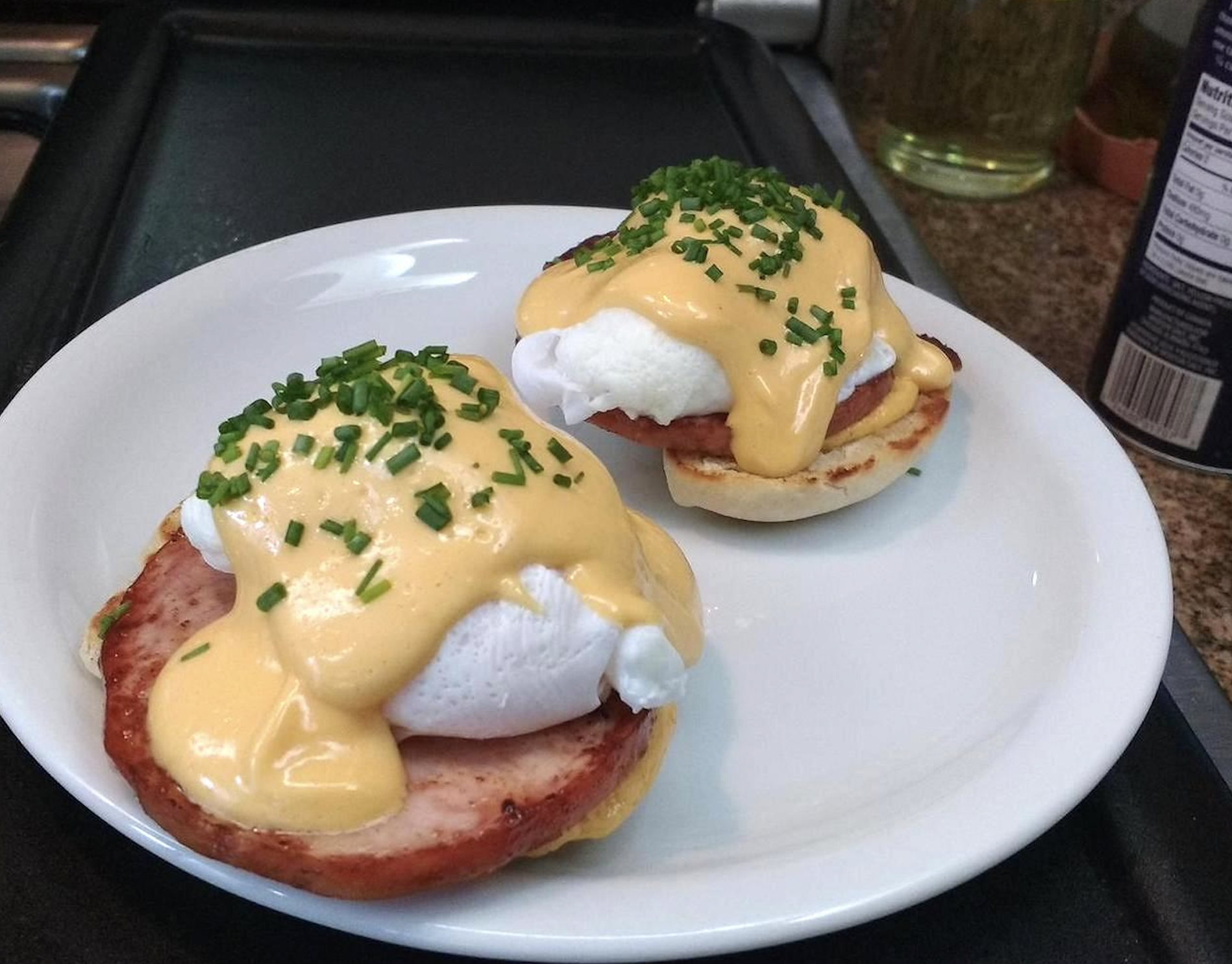 Two eggs Benedict on a white plate, topped with hollandaise sauce and chopped chives, sitting on slices of ham and toasted English muffins.
