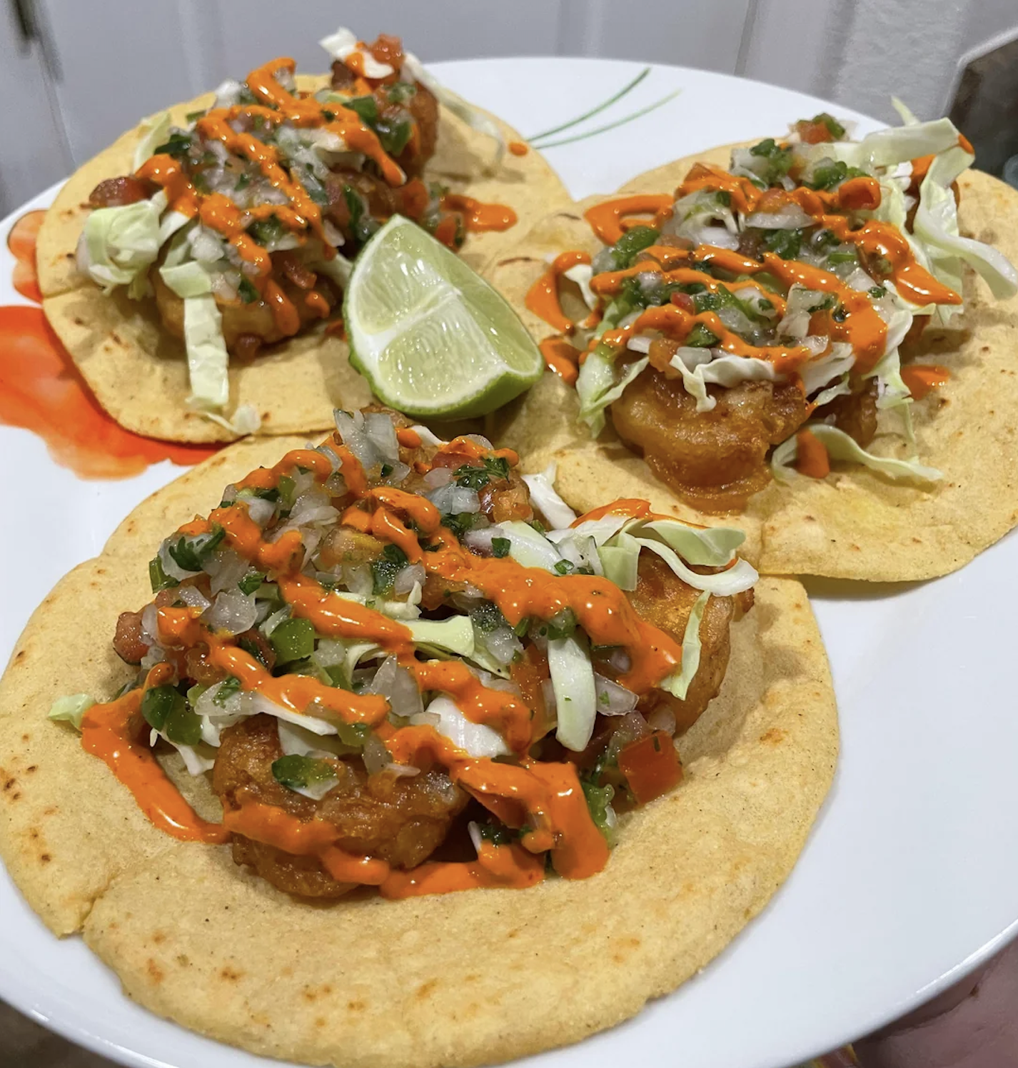 Three tacos on a plate, each topped with shrimp, chopped vegetables, shredded cabbage, and orange sauce, served with a lime wedge.