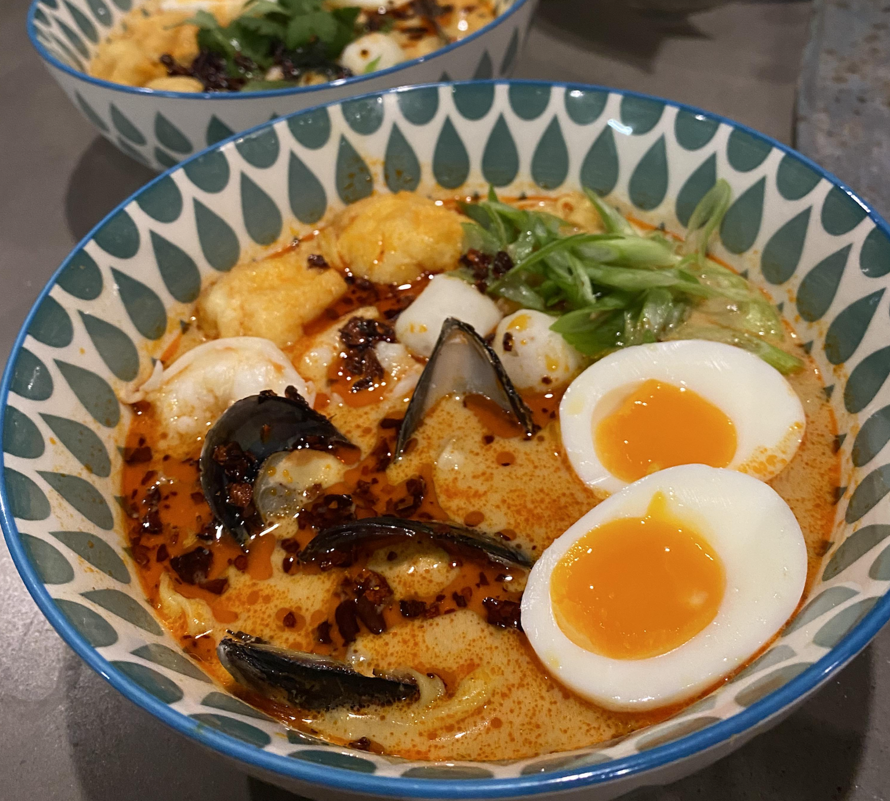A bowl of spicy seafood ramen with mussels, shrimp, soft-boiled egg halves, green onions, and chili oil in a patterned bowl. Another similar bowl is blurred in the background.