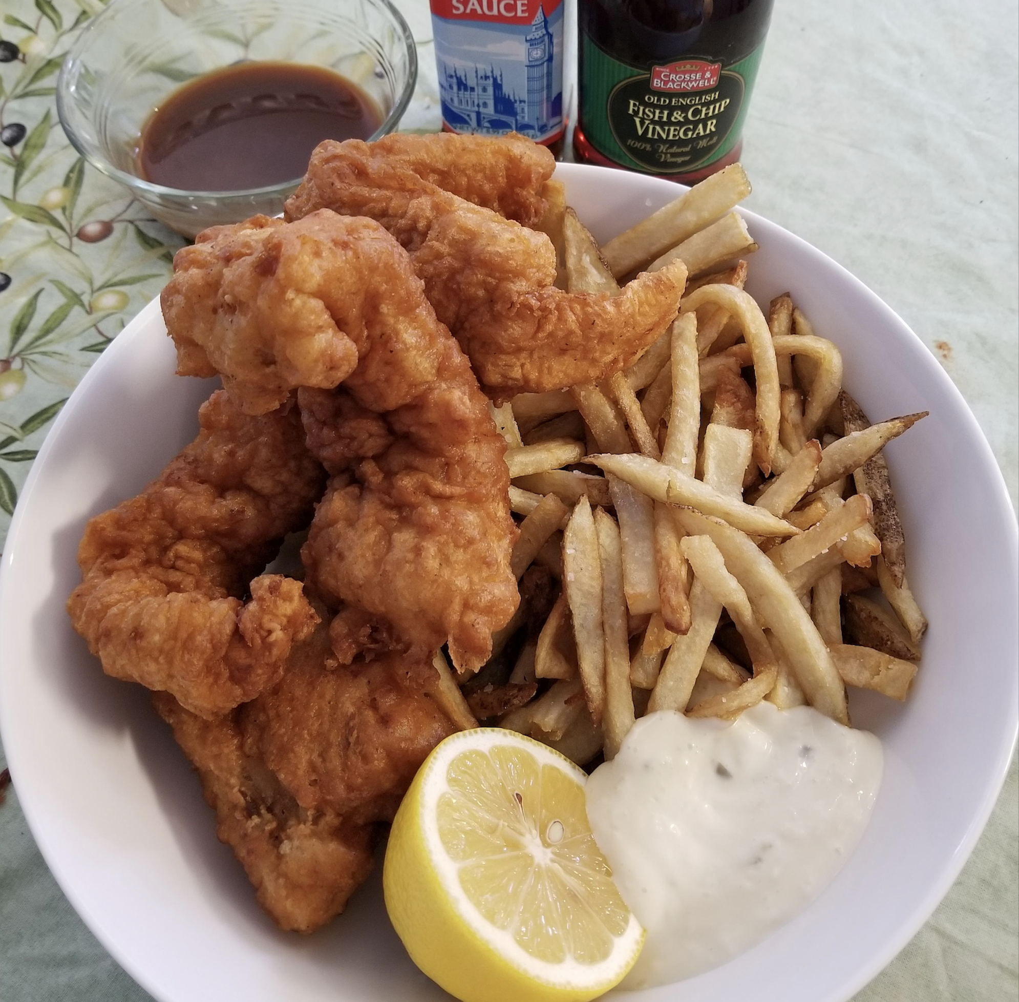 A bowl of crispy fried fish, golden French fries, tartar sauce, and a lemon wedge. In the background are bottles of malt vinegar, fish and chip sauce, and a small bowl of brown dipping sauce.