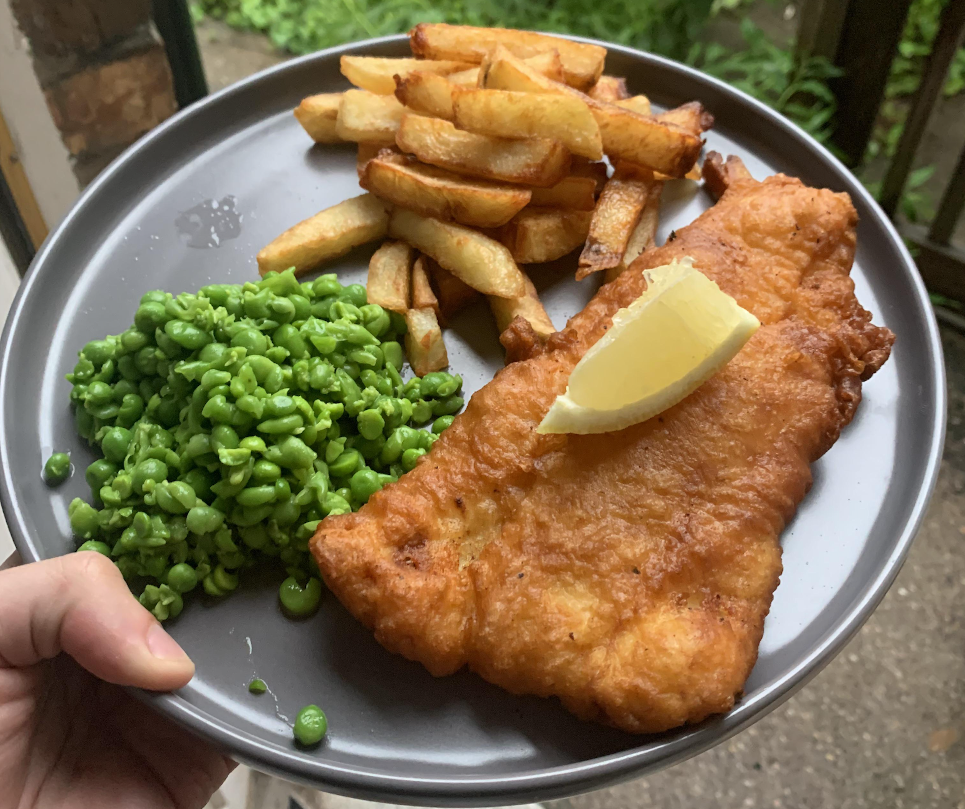 A plate with battered fried fish topped with a lemon wedge, a serving of thick golden fries, and a portion of mushy peas. The meal is held outdoors near a window with green plants visible in the background.
