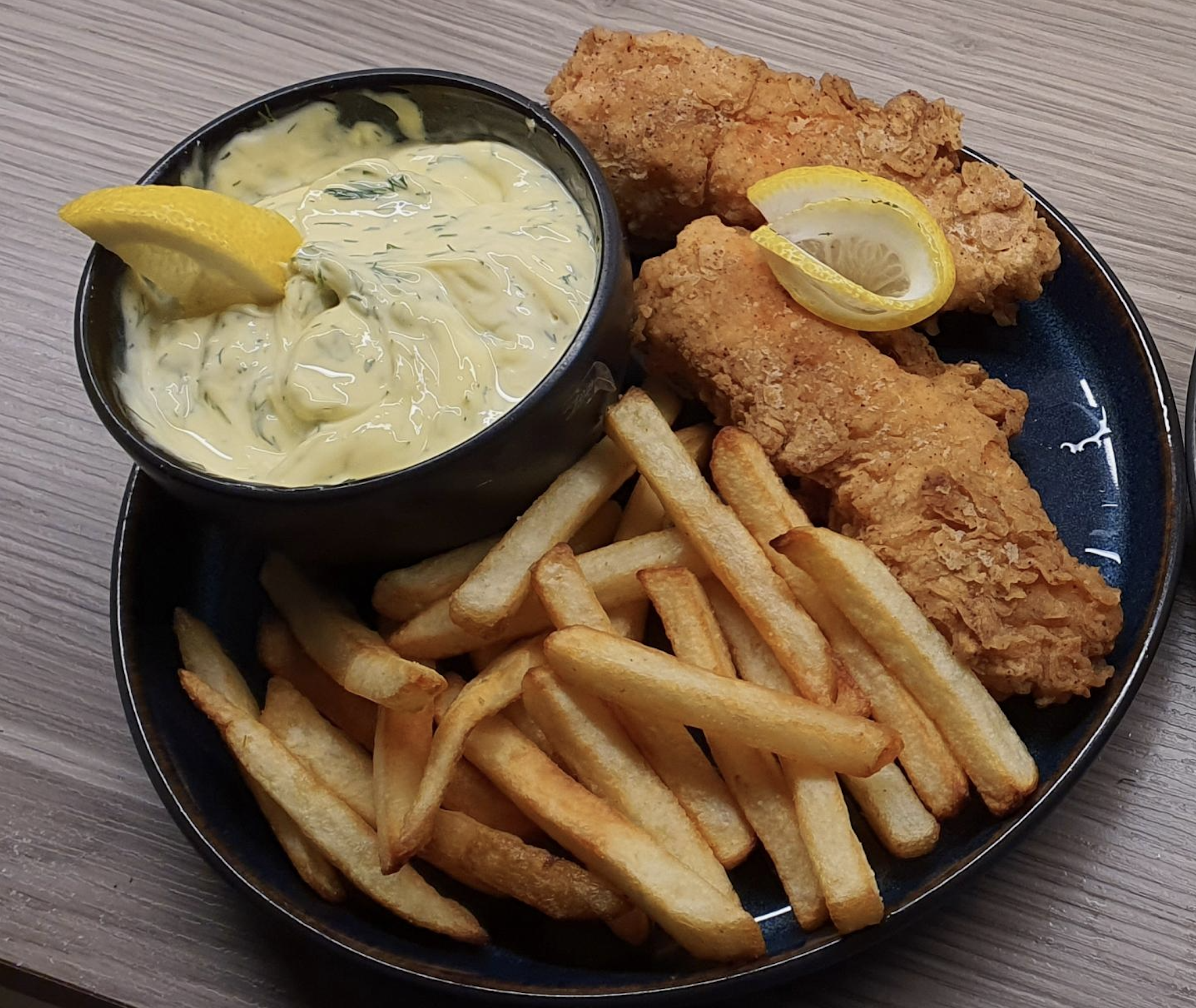 A plate of golden French fries and crispy fried fish fillets, garnished with a lemon wedge, served with a bowl of creamy tartar sauce topped with a lemon slice.