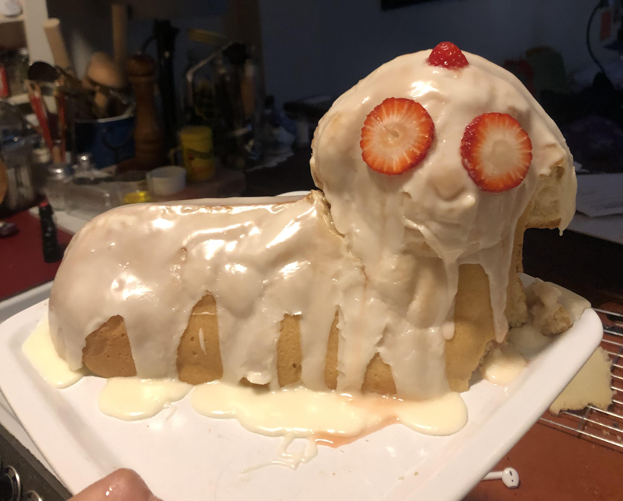 A loaf cake covered in white icing, shaped like a creature with strawberry slices for eyes and a nose, with icing dripping down to resemble a face.