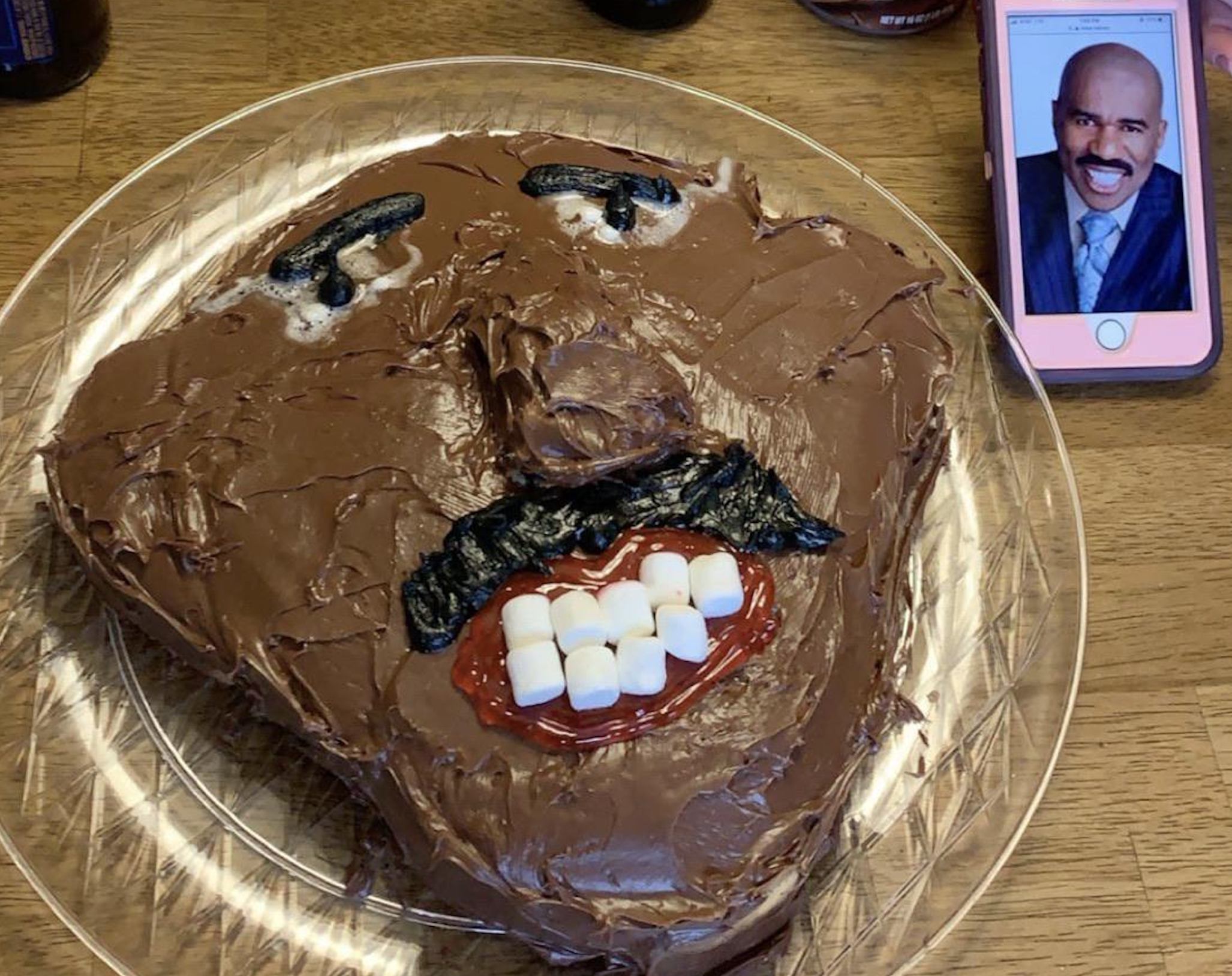 A cake decorated to look like a man’s face with chocolate frosting, marshmallows for teeth, and black icing for a mustache and eyebrows. A phone beside it displays a photo of a man in a suit for reference.