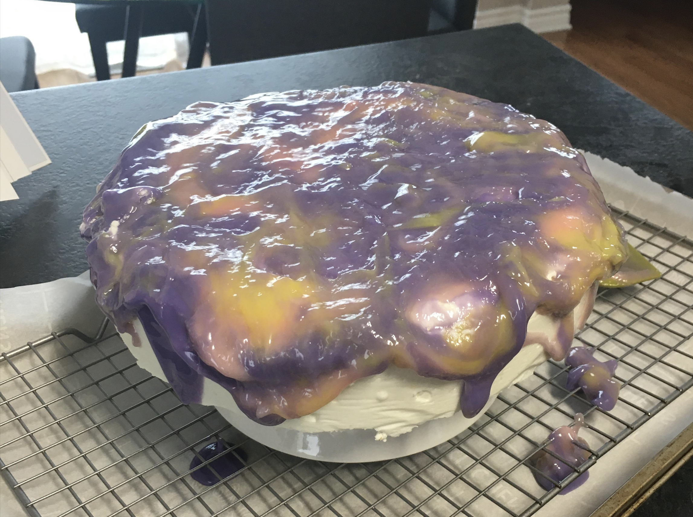 A round cake with white icing is topped with thick, glossy purple and yellow glaze that drips over the sides, sitting on a wire rack lined with parchment paper.