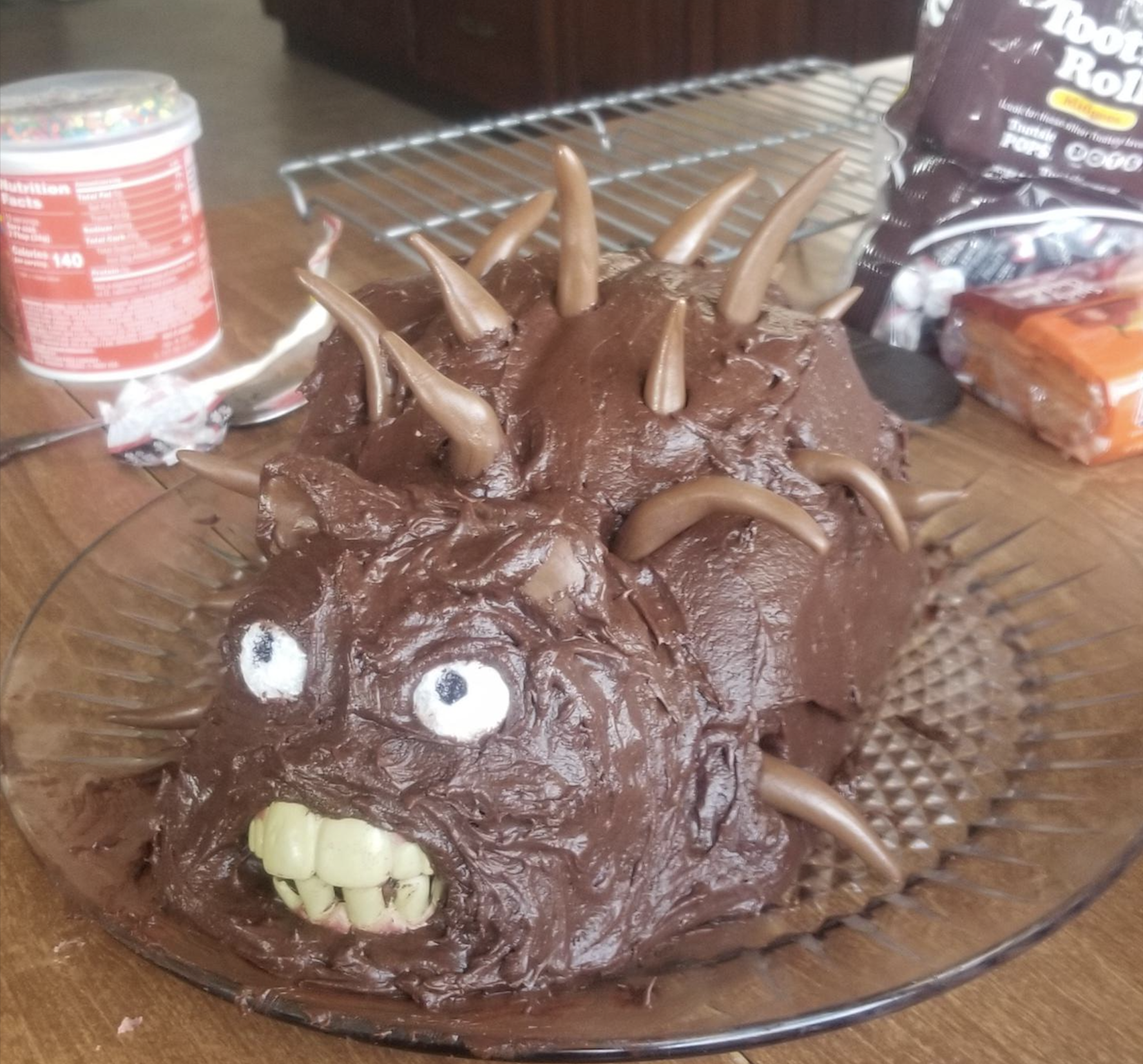 A homemade chocolate cake shaped like a monster, with chocolate spikes, large white eyes, and green candy teeth, sits on a glass plate. Baking supplies are scattered in the background.
