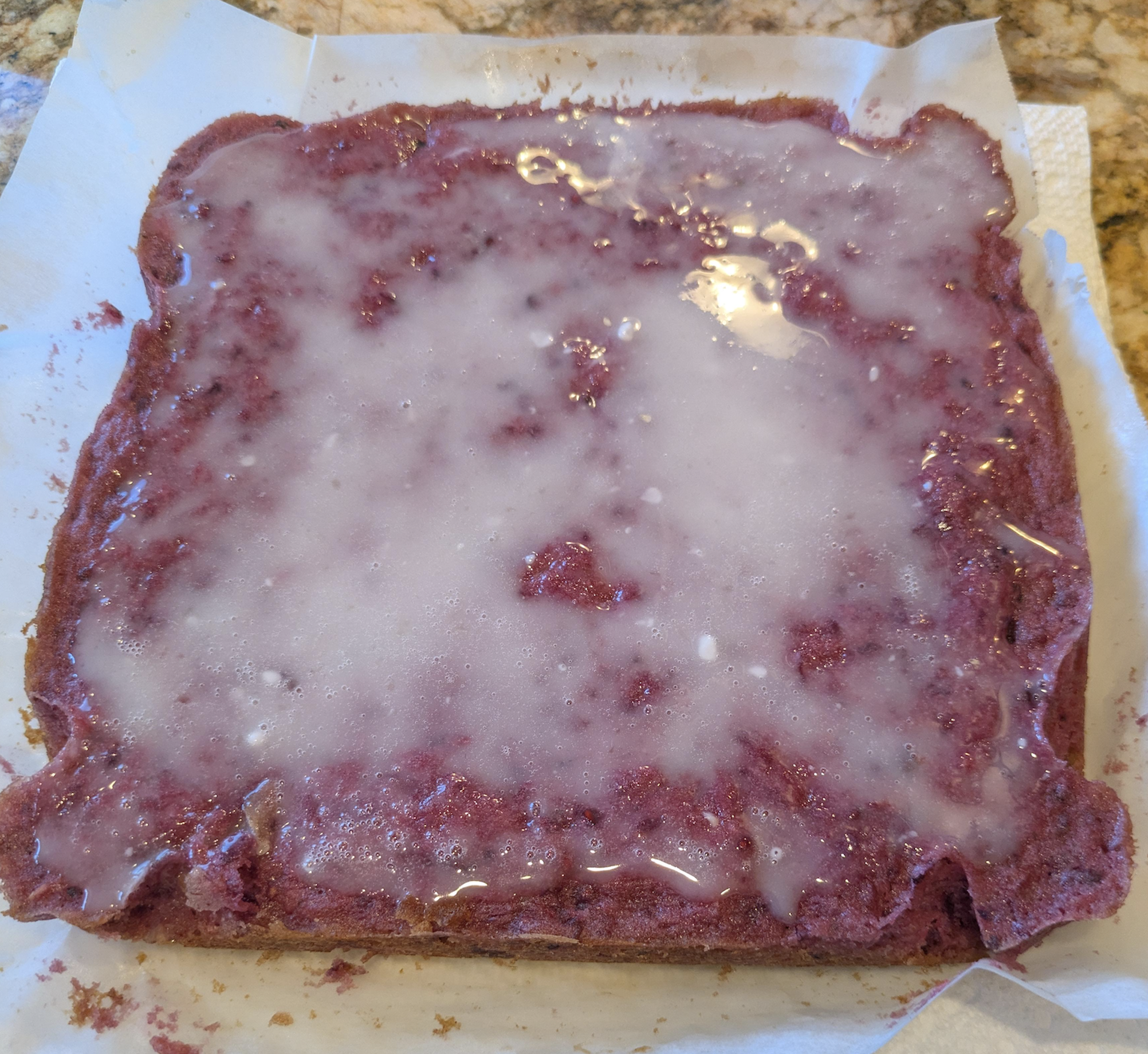 A square dessert with a purple base and a shiny white glaze on top, sitting on parchment paper on a kitchen counter. The edges are uneven and the glaze has small bubbles.