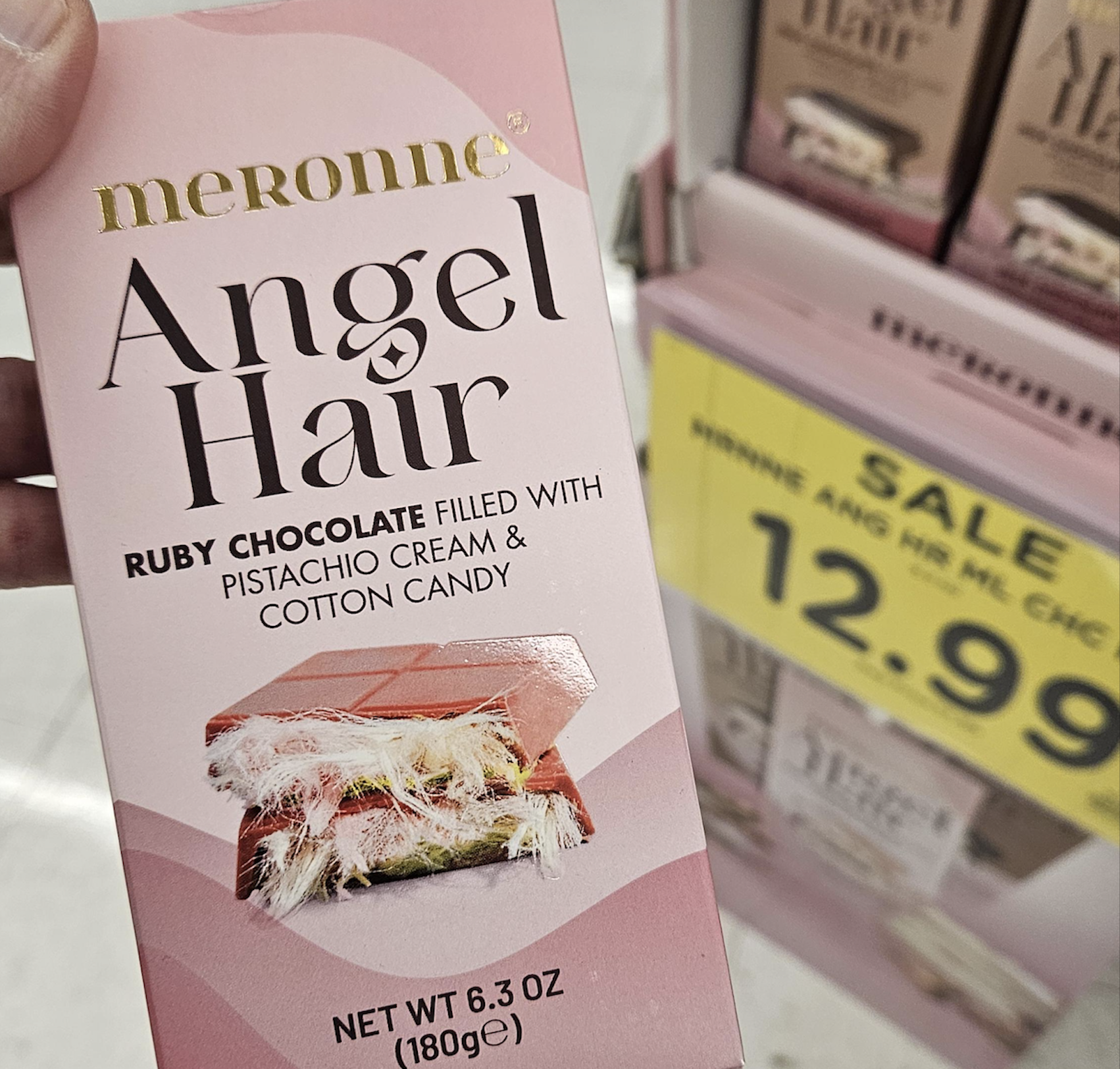 A hand holds a box of Meronne Angel Hair ruby chocolate with pistachio cream and cotton candy. A yellow sale sign beside a display shows the price as $12.99.