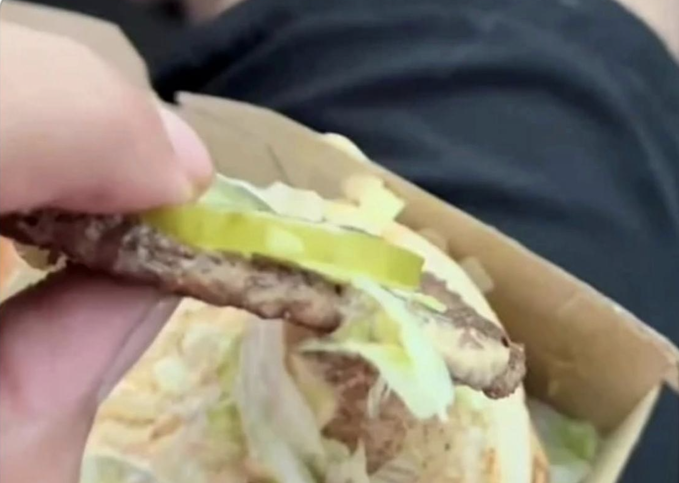A hand holds a thin burger patty with lettuce and two pickle slices, lifted above an open wrapper with more lettuce visible below.