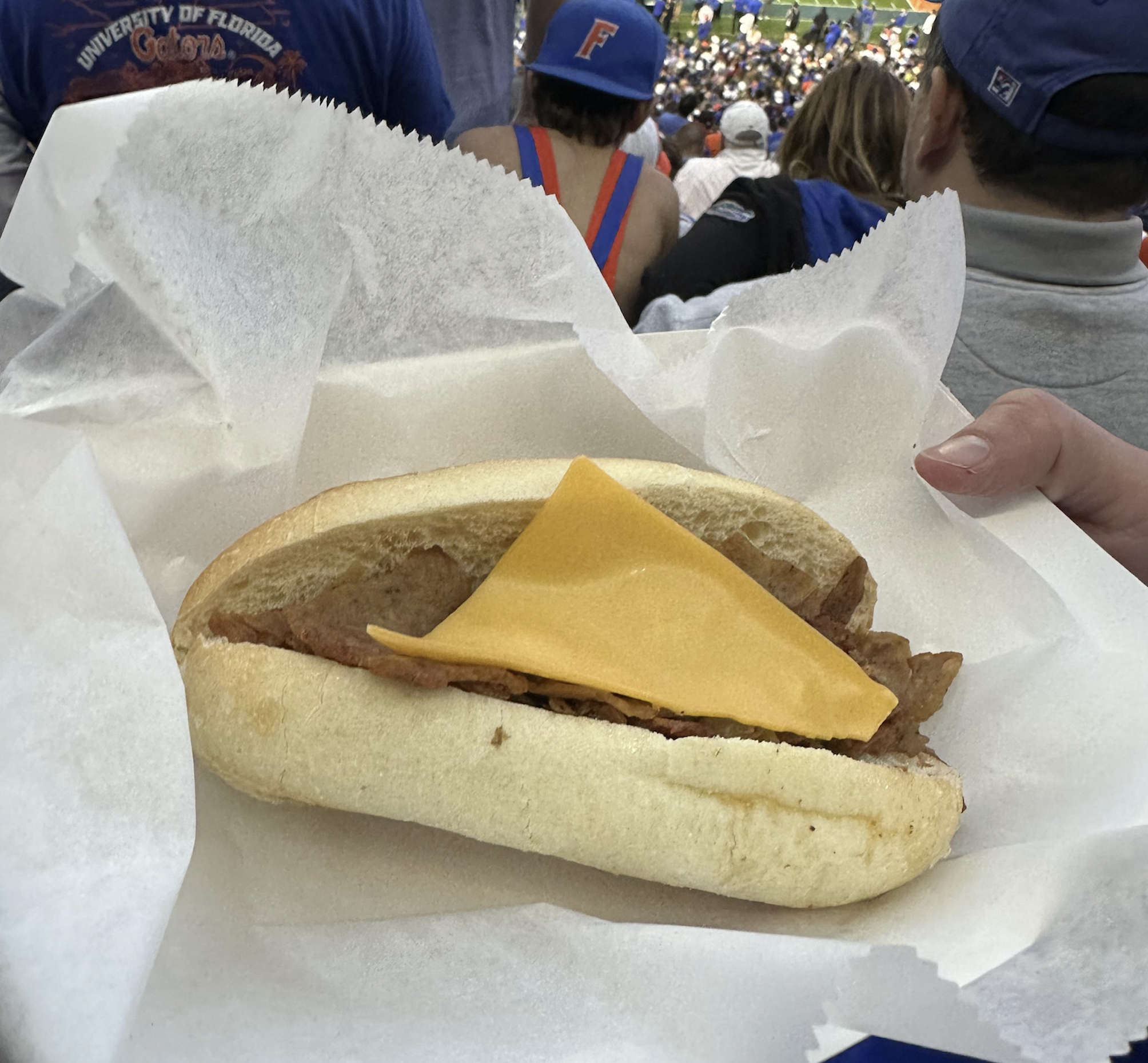 A person holds a sandwich with sausage and a slice of cheddar cheese on a white bun, wrapped in paper, at a crowded stadium event.